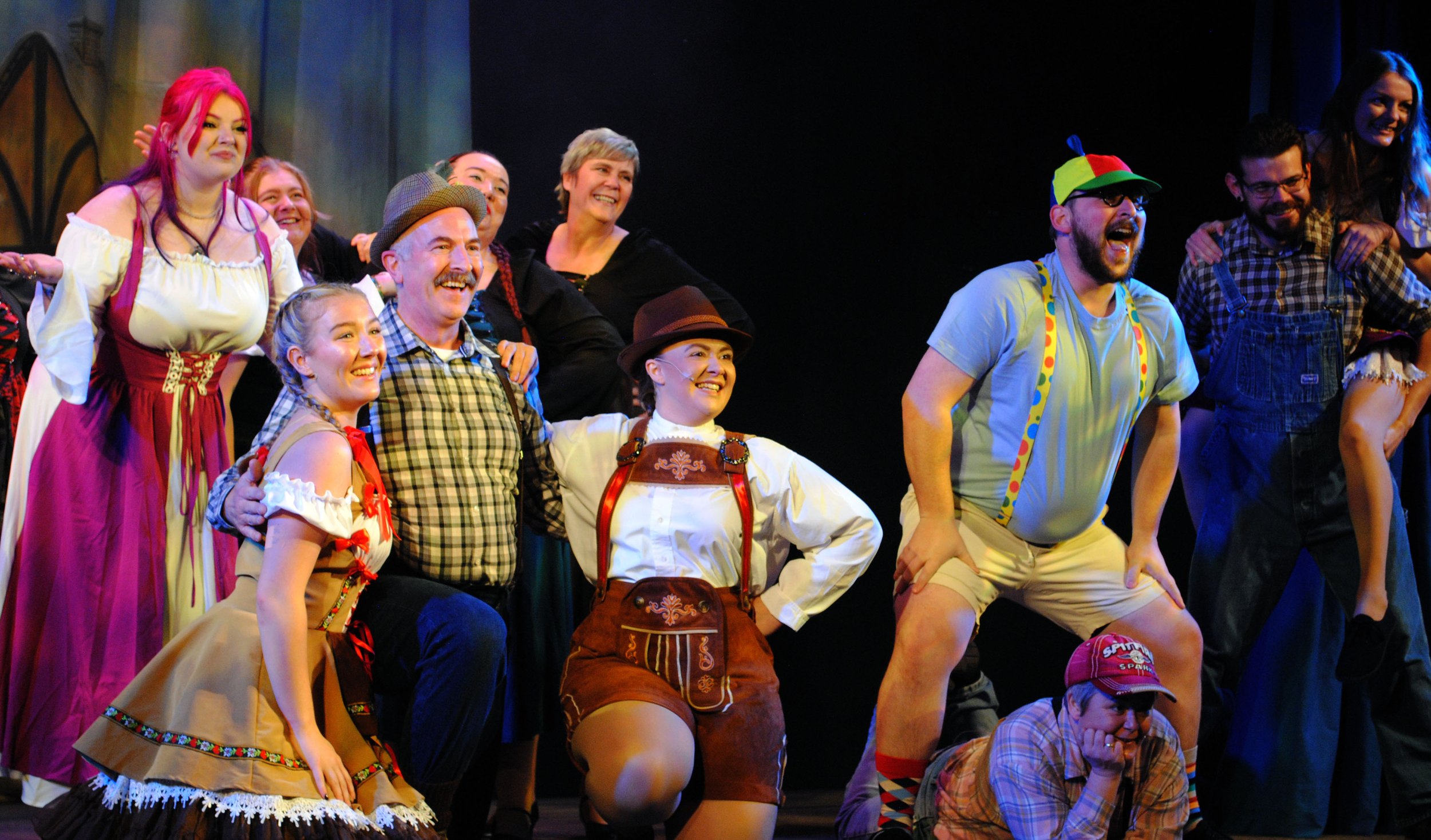 The cast of a theatrical play dressed in colorful costumes, smiling and posing on stage for a group photo.