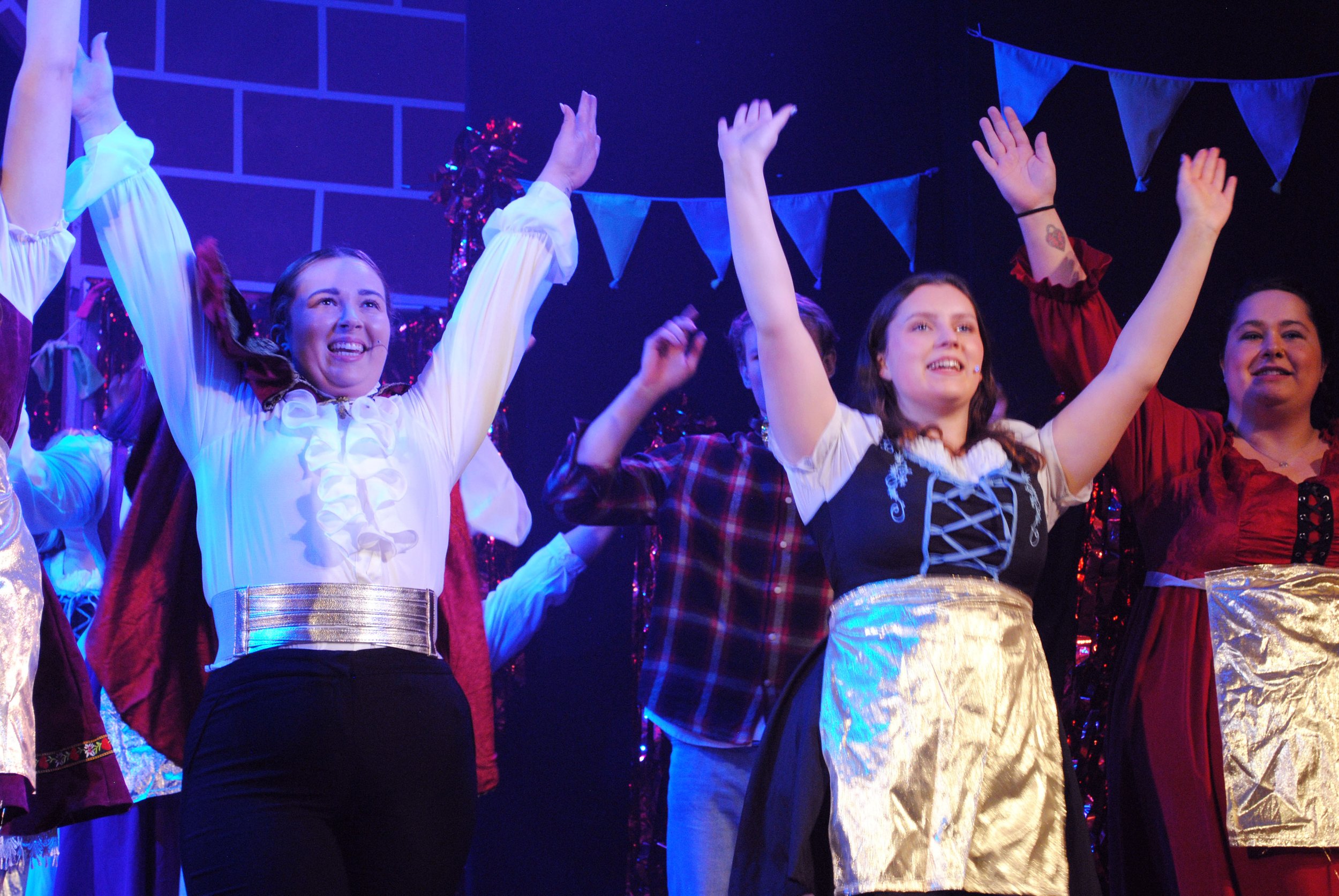 Group of women dressed in costumes on stage, raising their hands and smiling, with blue bunting and Christmas decorations in the background.