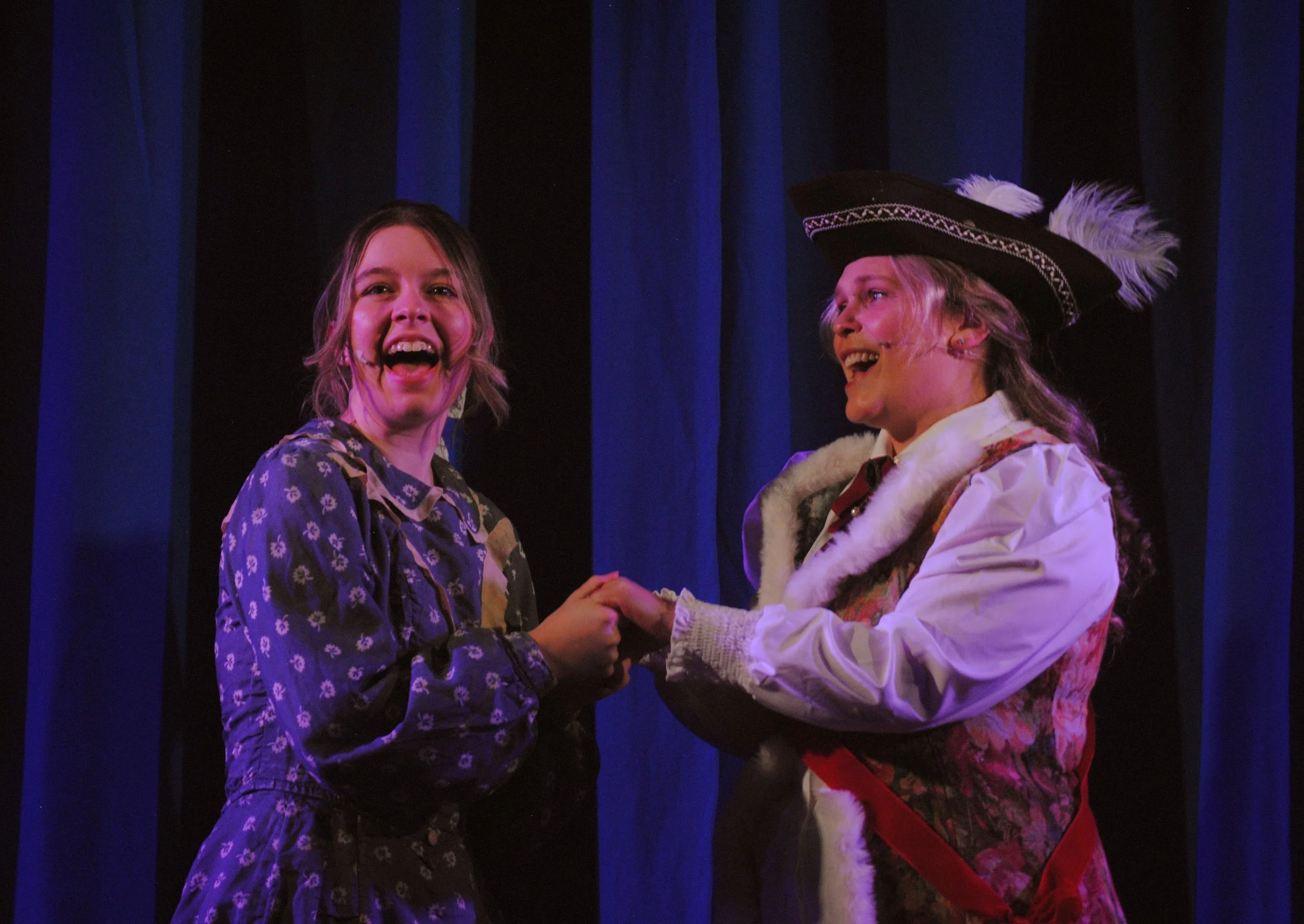Two women on stage, one dressed in a floral patterned dress and the other in a historic costume with a feathered hat, smiling and holding hands during a theatrical performance.
