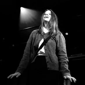 A woman smiling and looking upwards on stage with a spotlight shining down, in black and white.