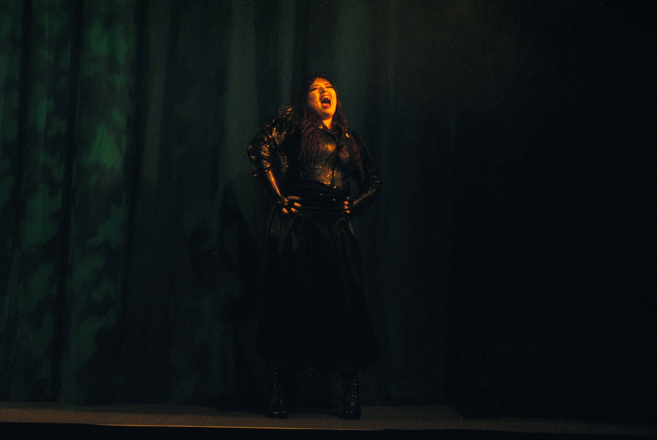 A woman on stage wearing a black leather outfit, with her mouth open and hands on her hips, performing under stage lighting with dark curtains in the background.