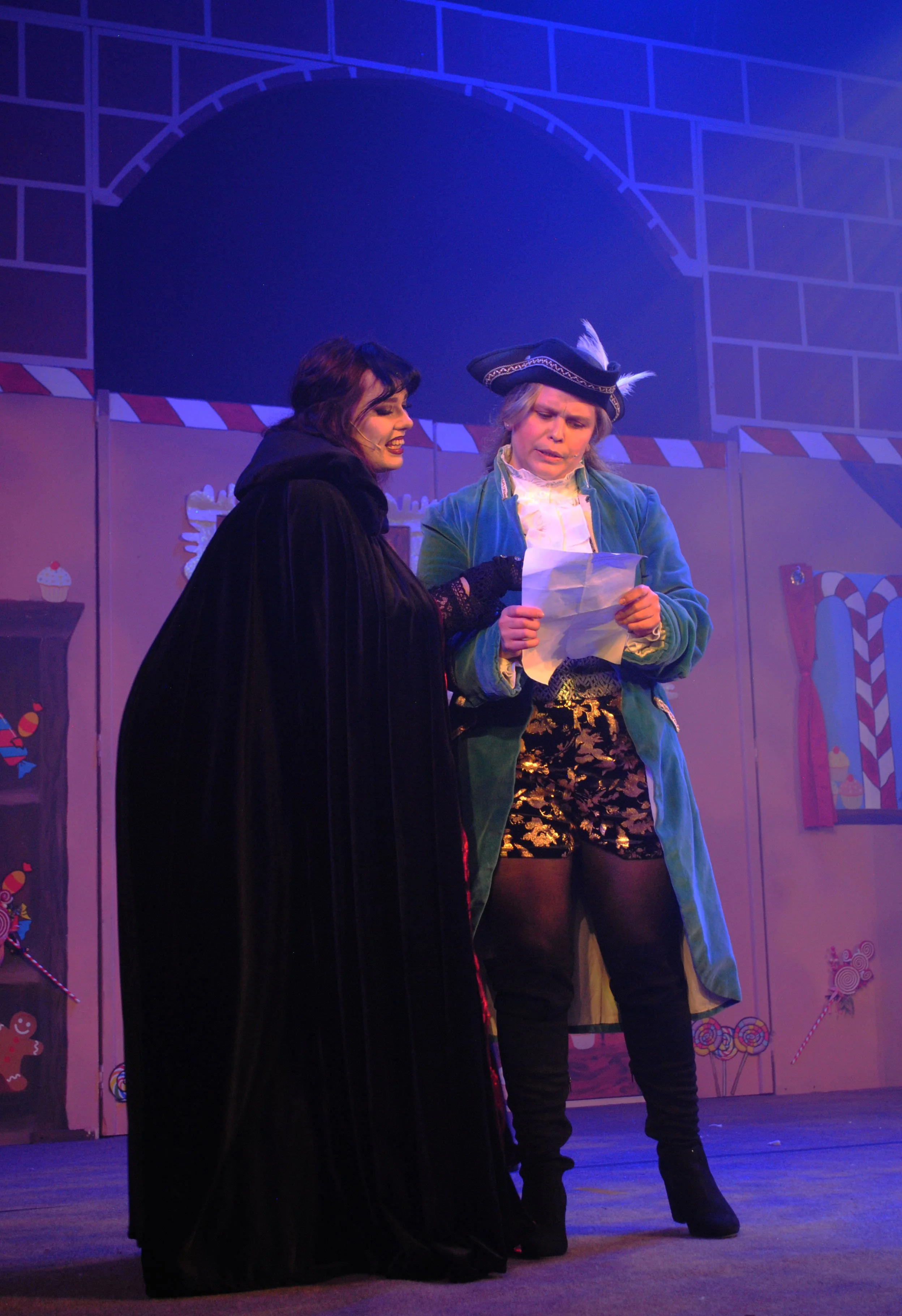 Two actors in costume on stage performing in a theatrical play, with festive holiday themed background decorations.