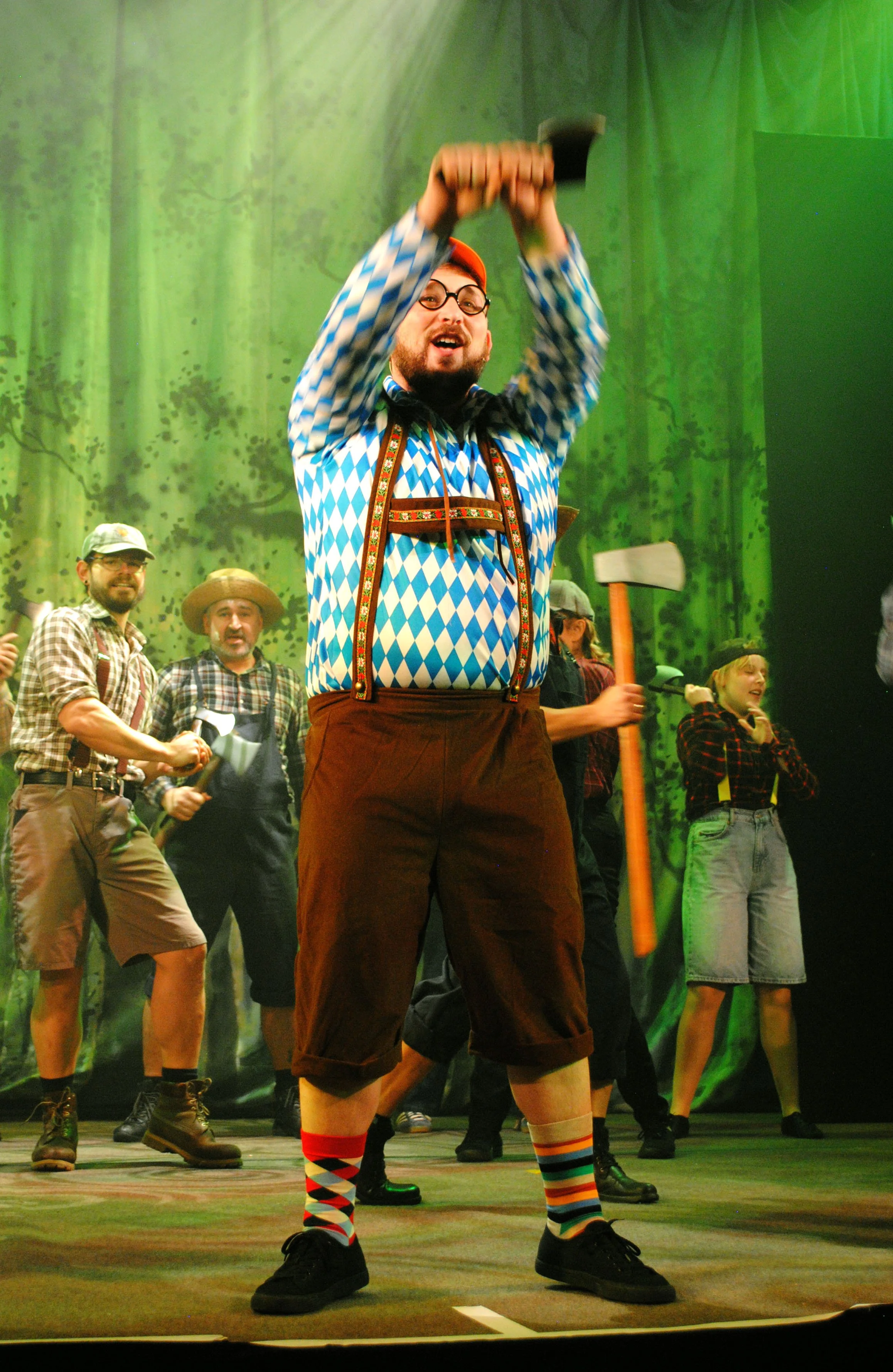 Group of people in costumes performing on stage, with the central figure dressed in Bavarian style, raising an axe in the air, and others holding axes and wearing plaid shirts, hats, and suspenders. The background features a green forest scene.