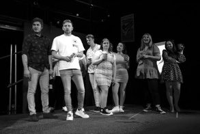 Group of six people standing on a stage, some holding microphones, in a black and white setting.