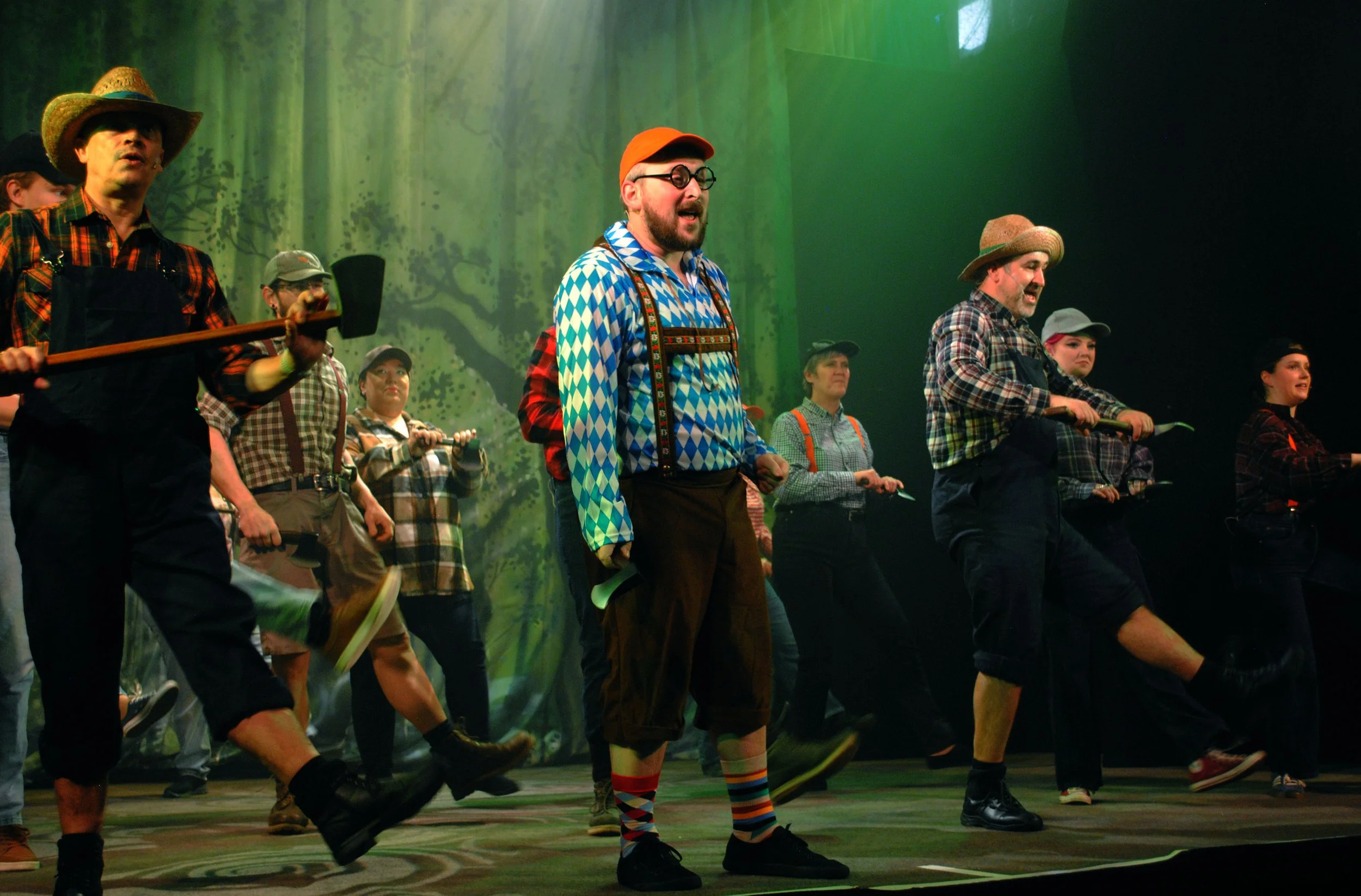 Actors on stage in a theatrical dance scene dressed in lumberjack and forest themed costumes, with a forest backdrop.