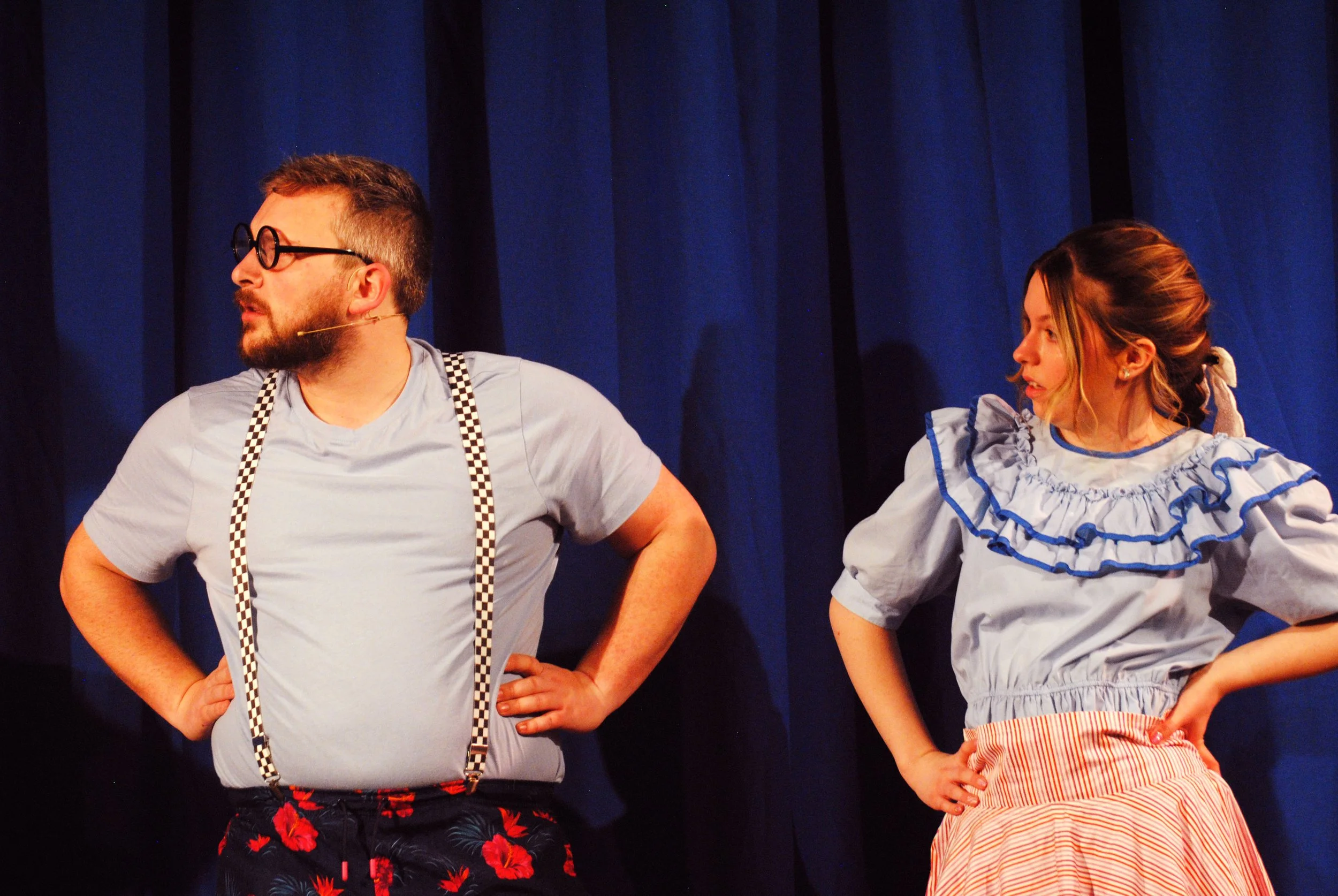 Two performers on stage with blue curtains in the background. The man on the left has a beard, glasses, and a light blue shirt with suspenders, and he is looking to his left. The woman on the right has light brown hair tied back, and is wearing a lig