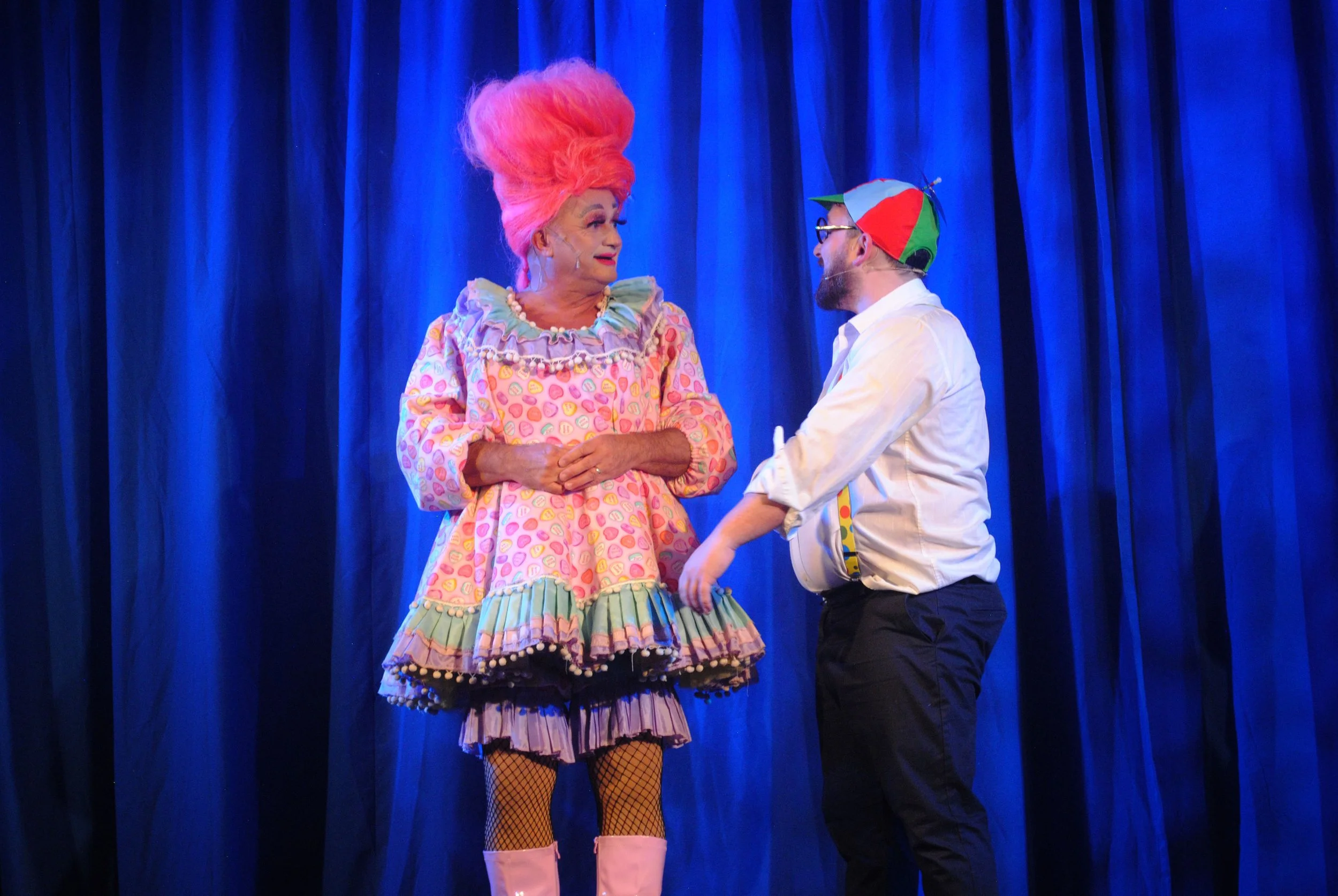 Two performers, a woman in colorful, whimsical clothing with bright pink hair and a man with a colorful cap, are on stage with a blue curtain backdrop, engaging in a theatrical act.