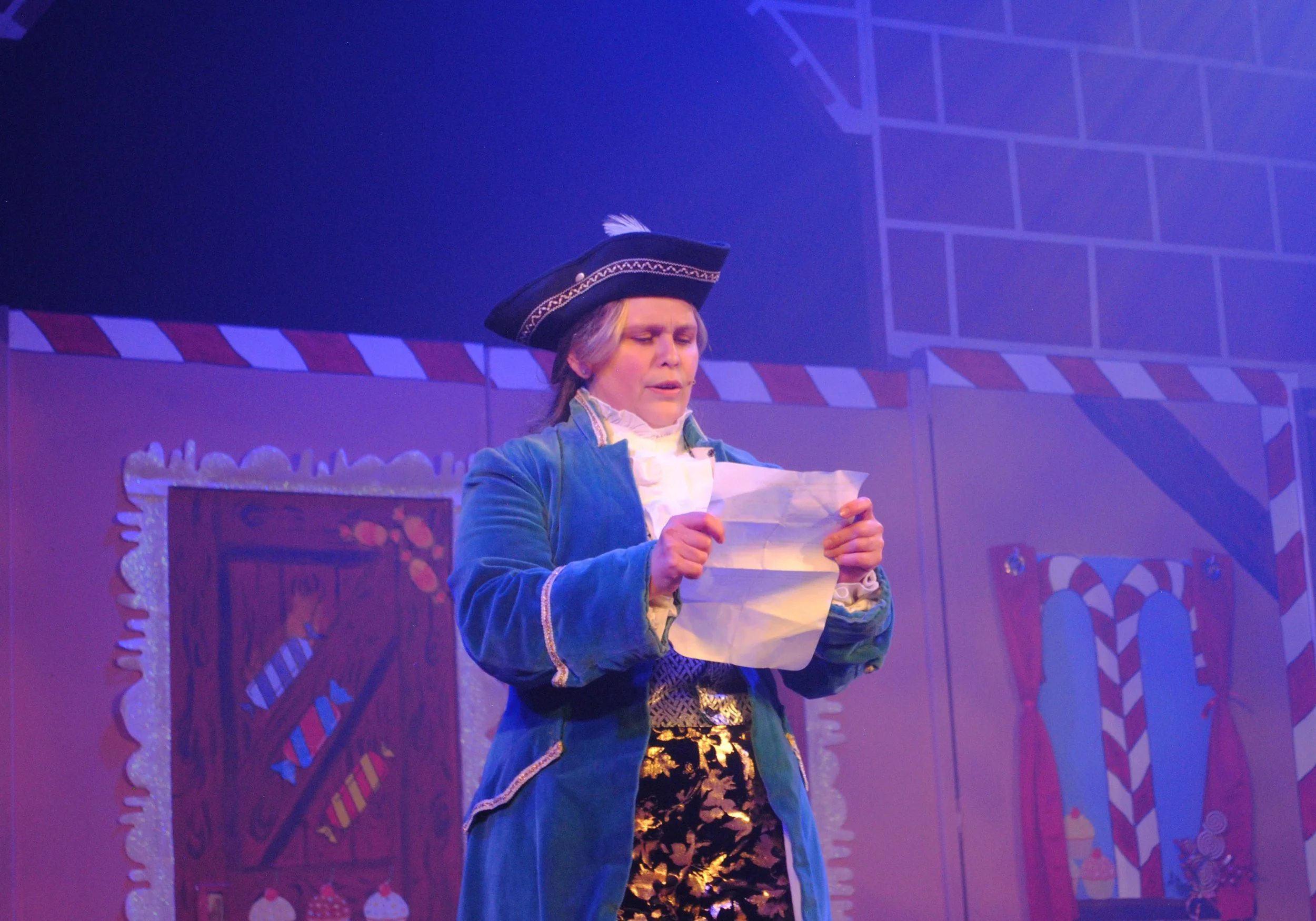 Person dressed in historical costume, reading a paper on stage with colorful, holiday-themed backdrop.