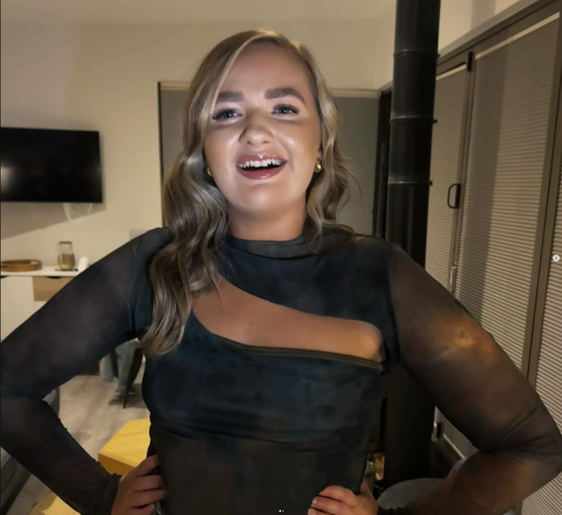 A woman with wavy hair and a black sheer top with cut-out detail, smiling and standing indoors in a modern living space.