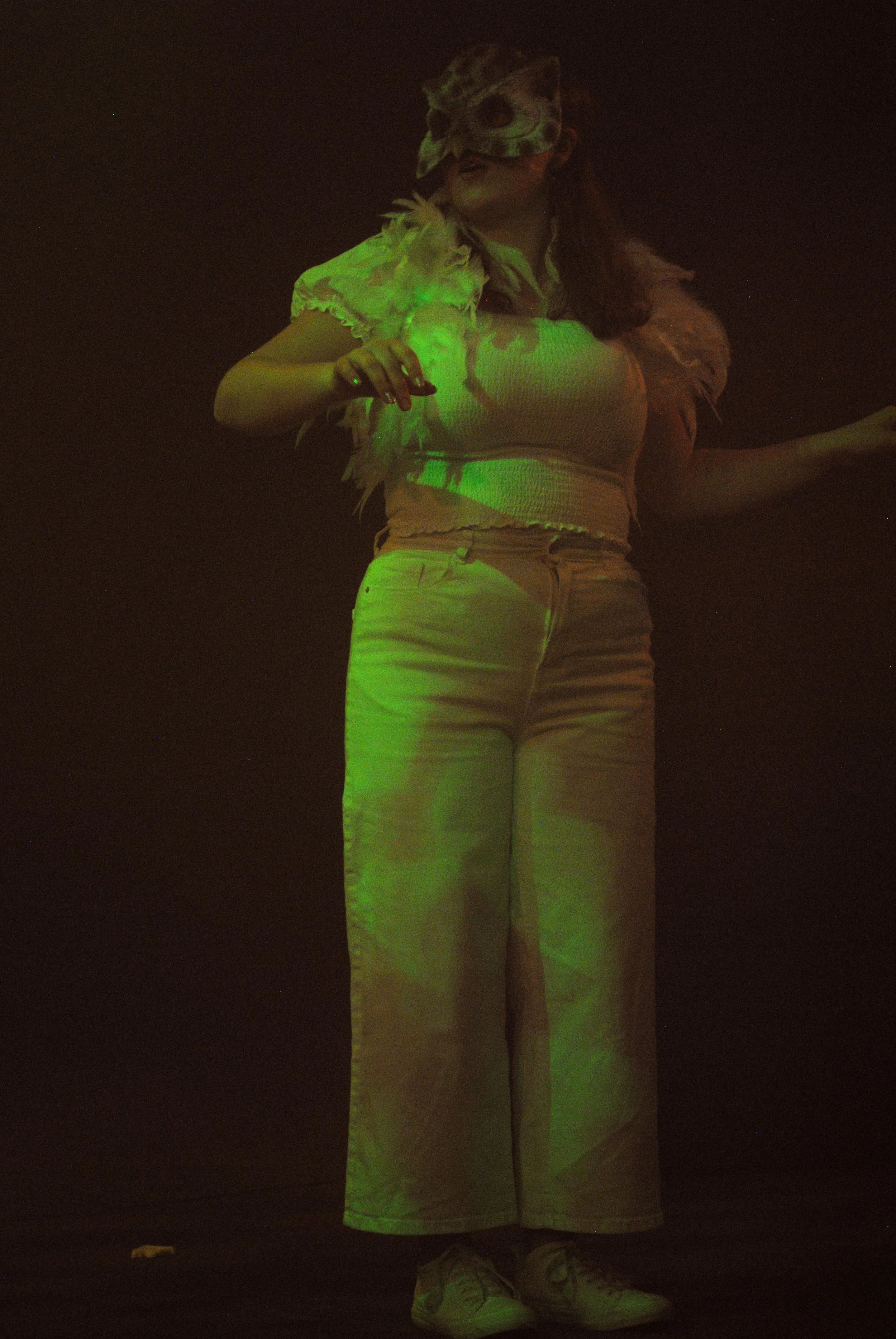 Person wearing a jaguar mask, green light cast, wearing a white and pink outfit, standing in a dark environment.