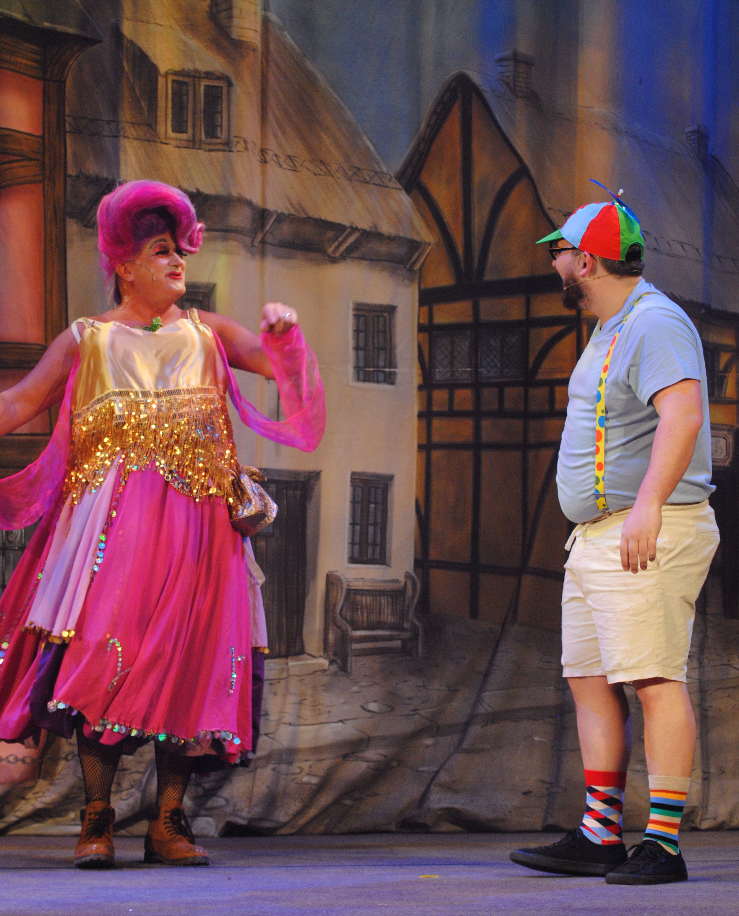 A woman with pink hair in a colorful costume and a man with a multicolored hat and socks on stage, with a painted backdrop of buildings.