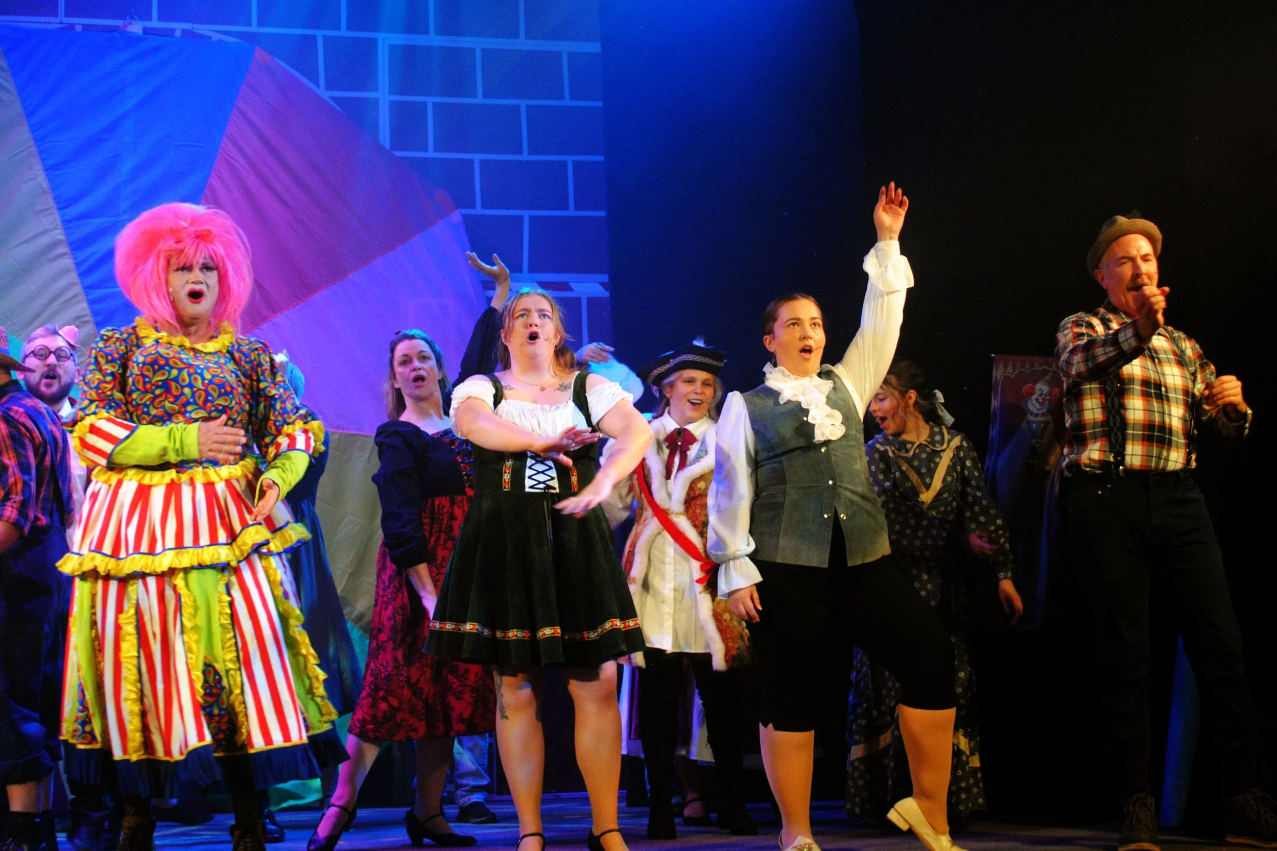 Cast of a theatrical performance dressed in colorful costumes, including a clown with pink hair, singing and performing on stage with a backdrop of a brightly colored house.