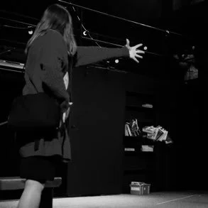 A woman in a long coat standing in front of a black box on a stage reaching out with her hand, in a black and white setting.