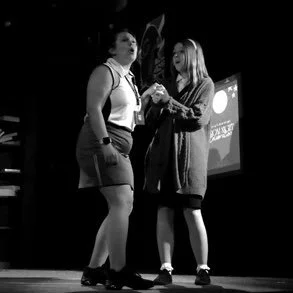 Two women standing on a stage, engaging in a conversation or interview, with one woman holding a microphone.