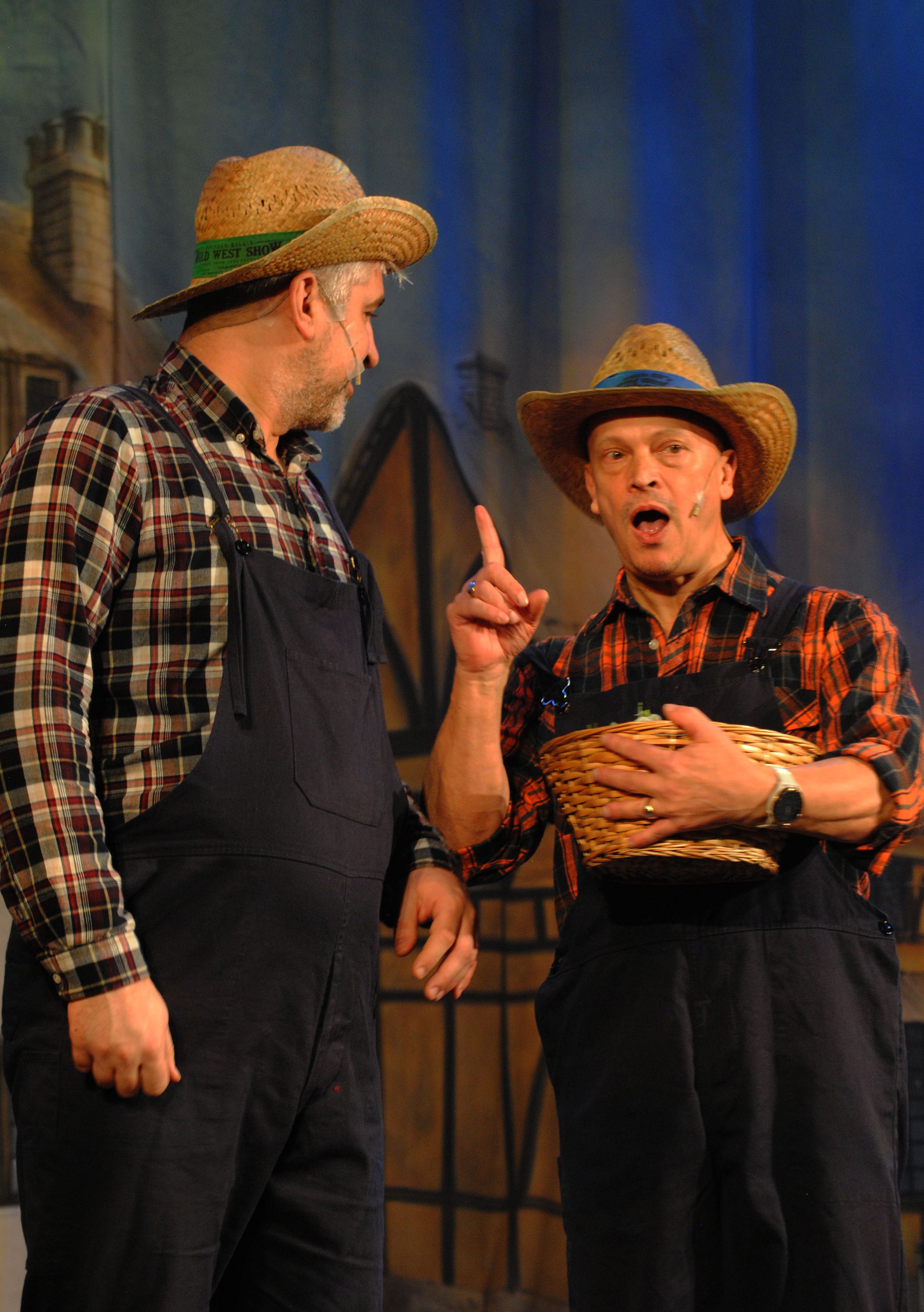 Two men dressed as farmers or workers, wearing straw hats and plaid shirts, are on stage with a painted backdrop of a building and sky. One man is gesturing with his finger while holding a basket, and the other is listening.