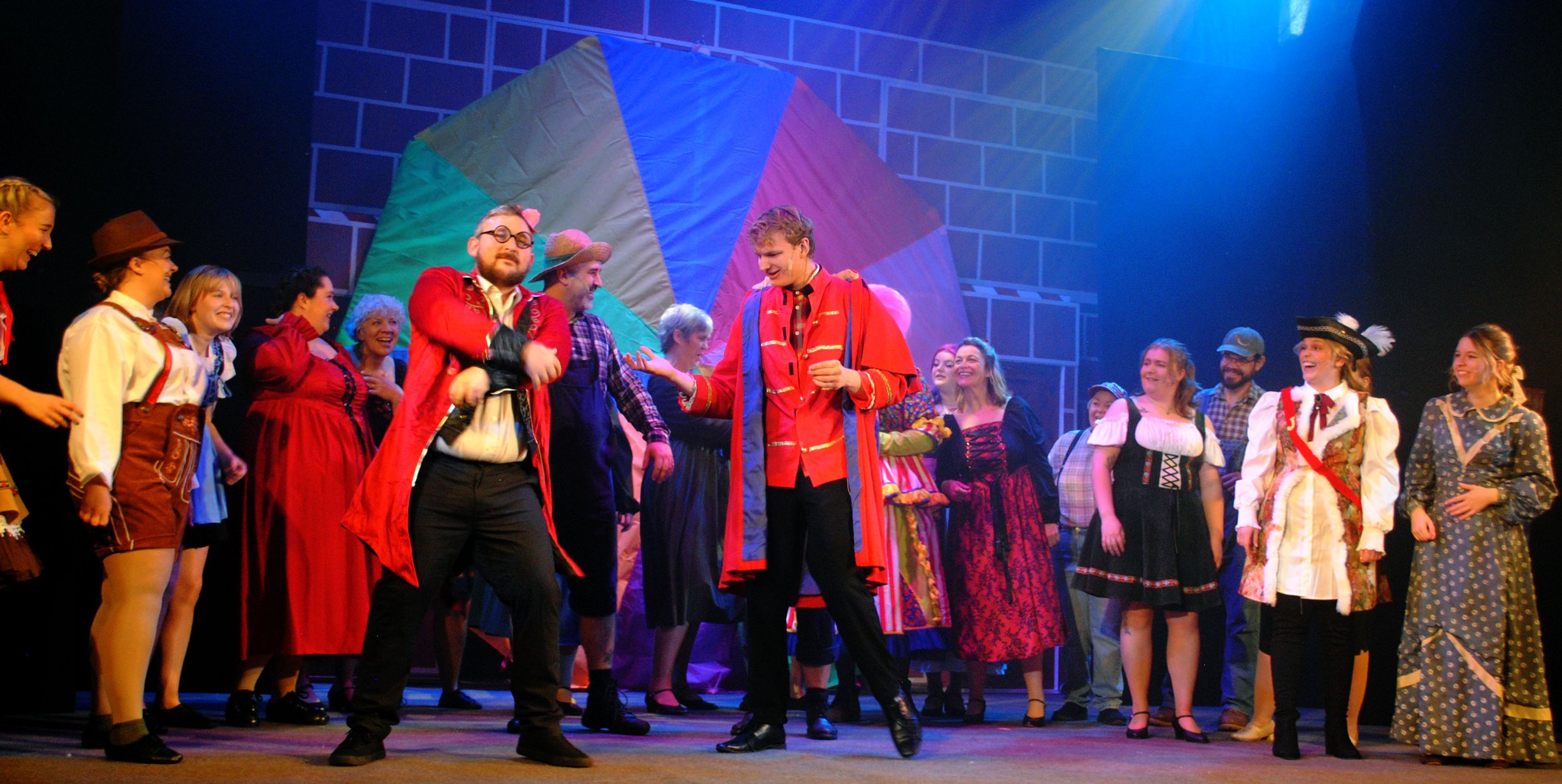 The cast of a theatrical play dressed in colorful costumes on stage, with some wearing historical attire, smiling and engaging in a lively scene with a large multicolored balloon in the background.