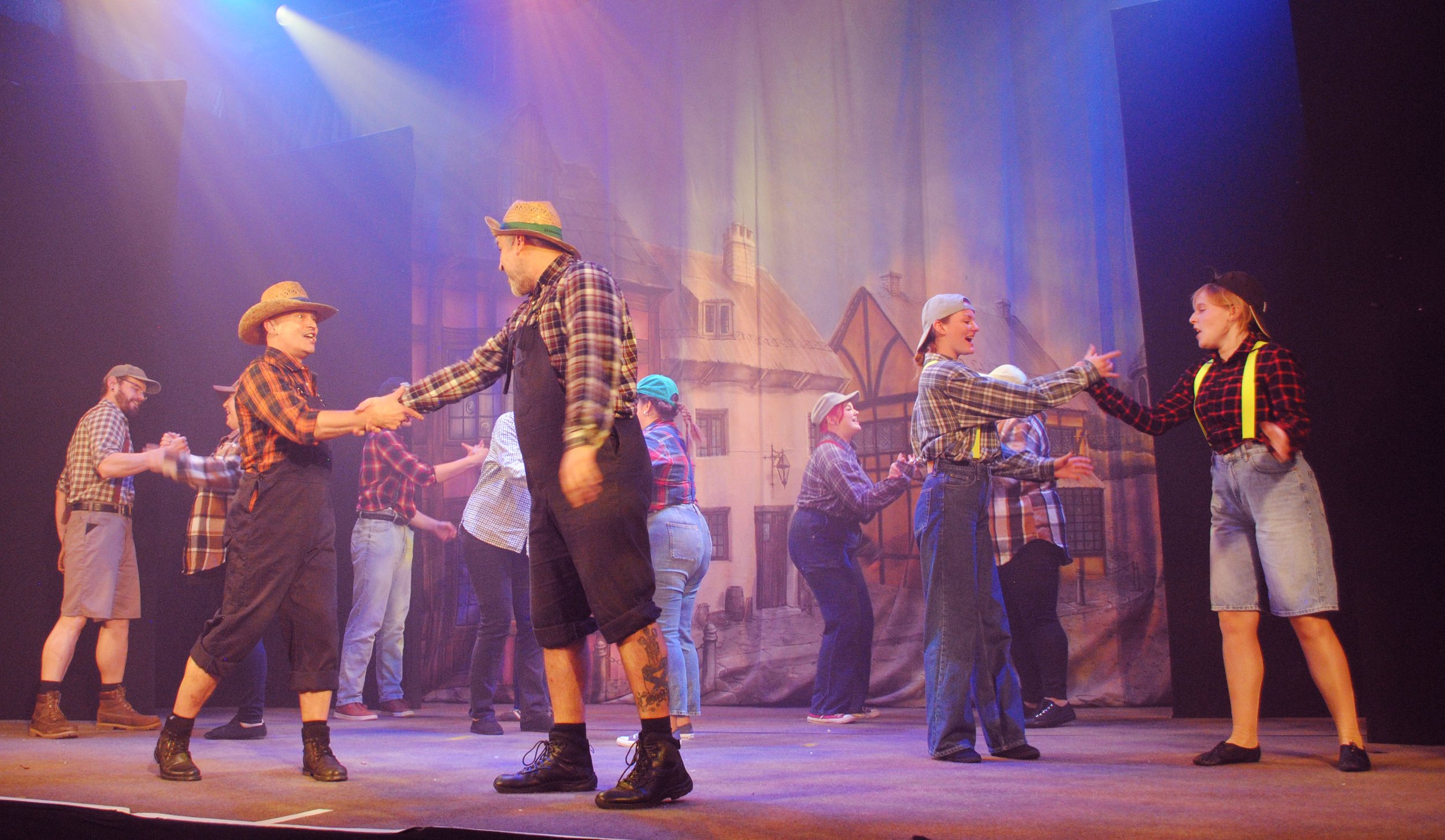People dressed as farmers on stage, shaking hands and dancing, with a painted backdrop of buildings and houses.