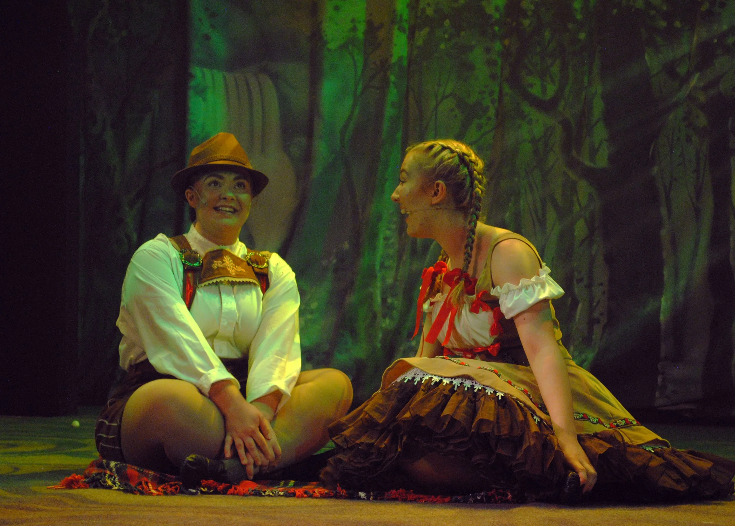 Two women in costume on stage, surrounded by a forest backdrop. One woman is wearing a hat and traditional clothing, sitting cross-legged with a cheerful expression. The other woman has braided hair and a colorful dress, also sitting on the floor, en
