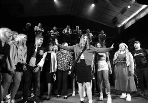 A group of people performing or rehearsing on stage with a woman in the center leading and others watching or participating.