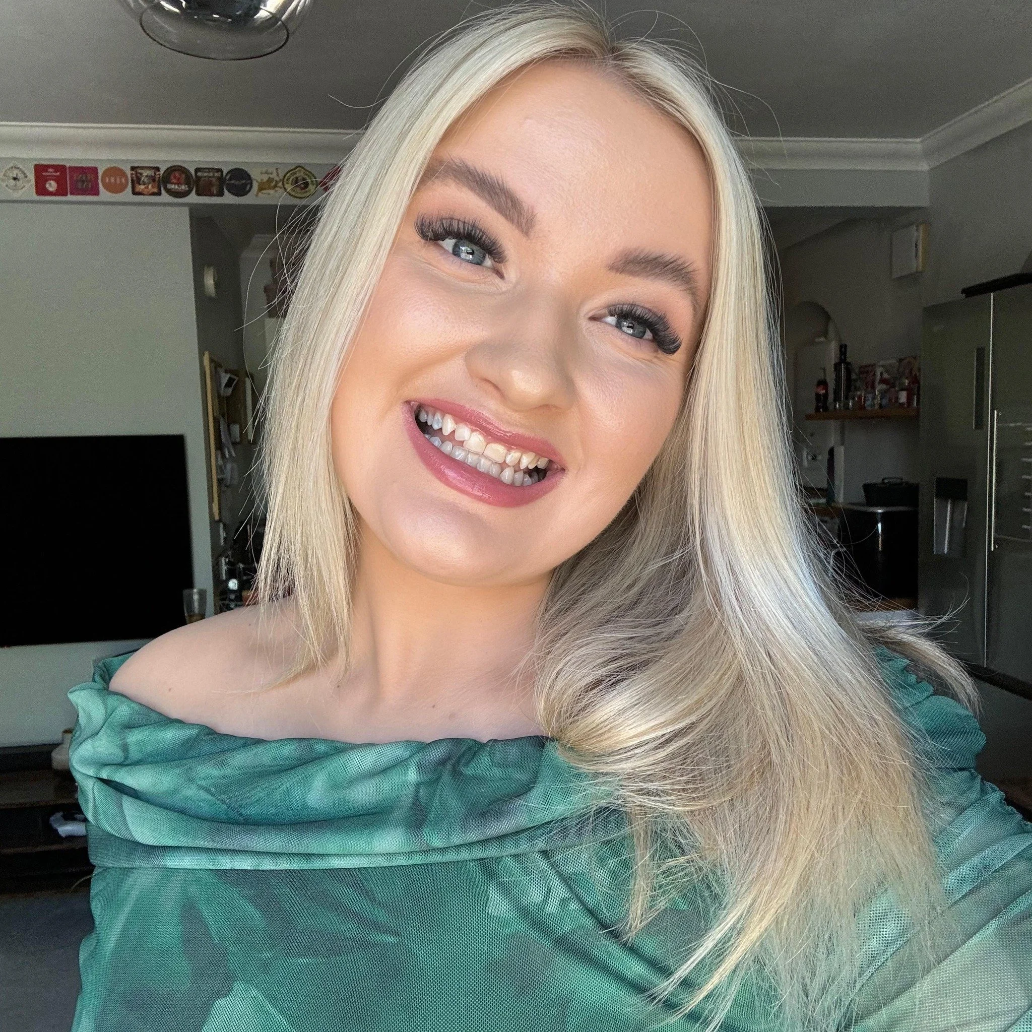 A young woman with blonde hair, blue eyes, and makeup, smiling and wearing a green off-shoulder top inside a home.