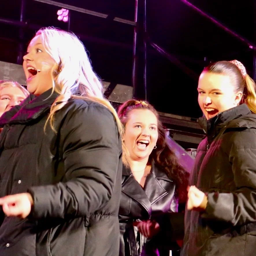 Four women on stage, smiling and reacting excitedly, dressed in winter jackets under bright stage lights.