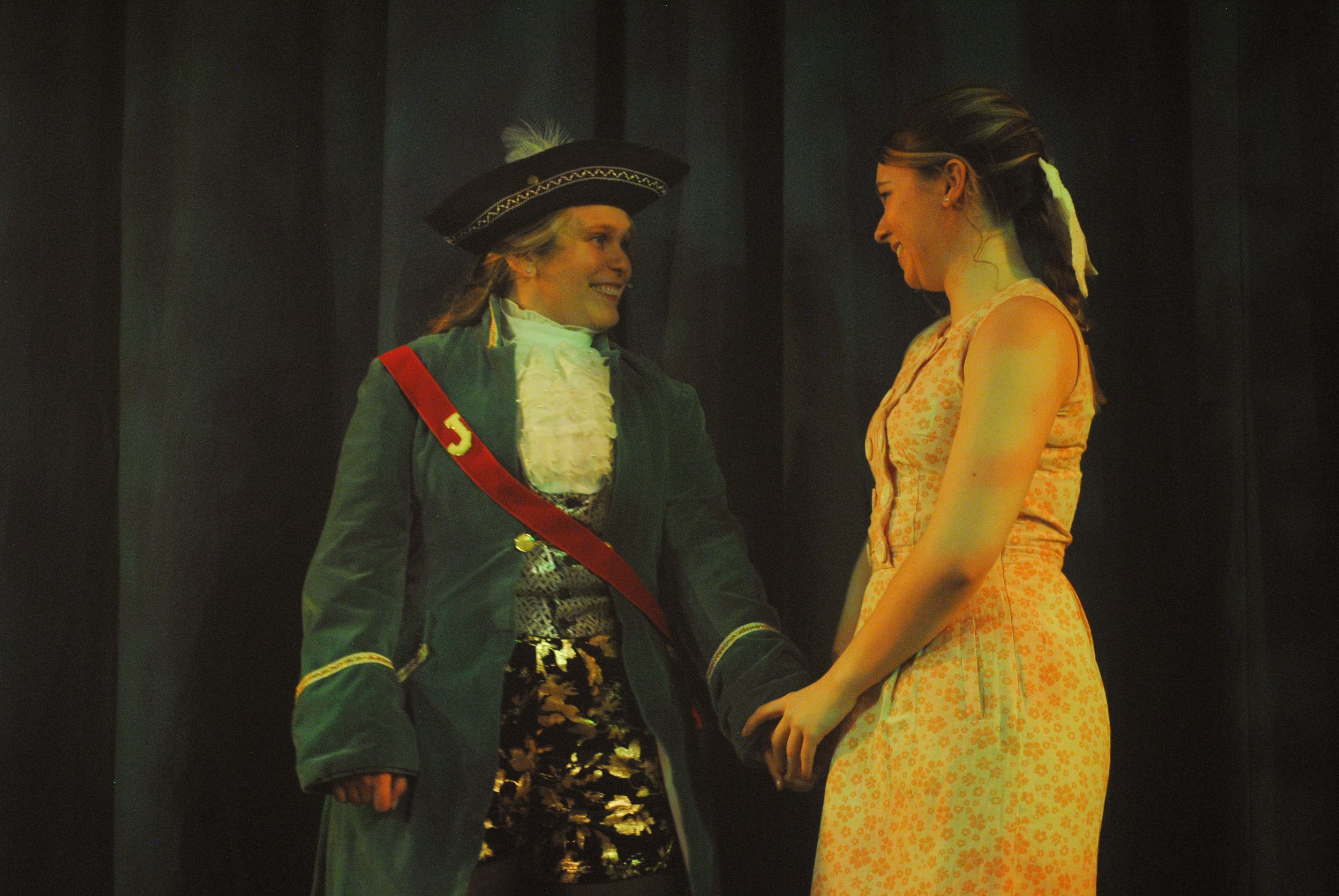 Two women dressed in period costumes, holding hands and looking at each other on stage, suggesting a theatrical performance.