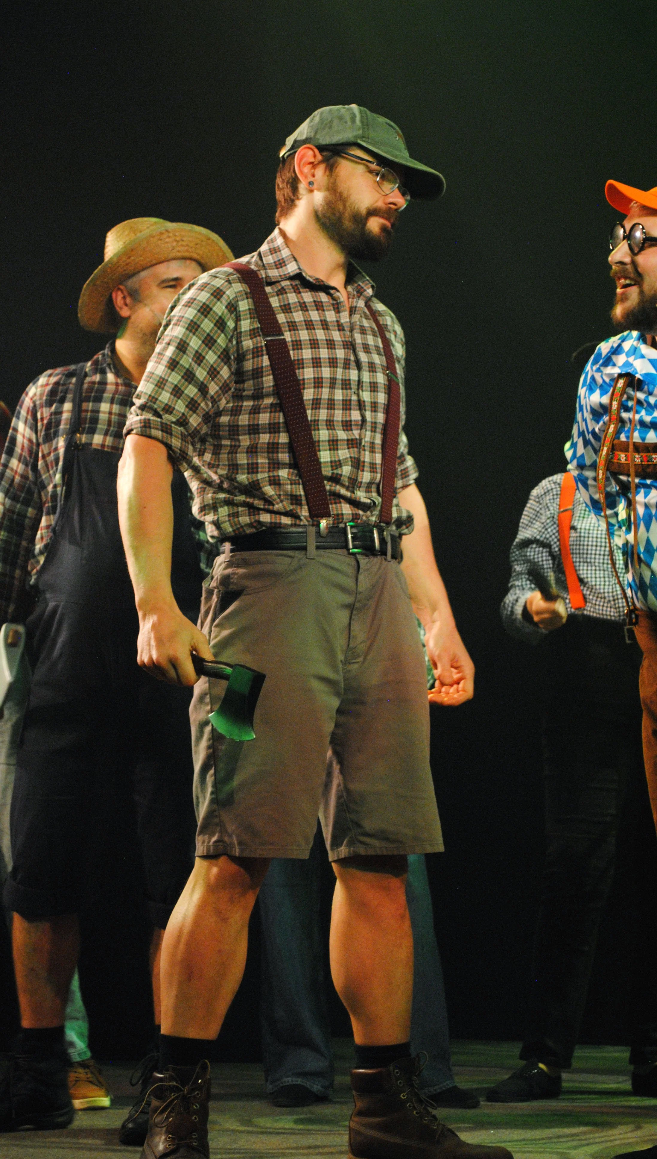 A man wearing a plaid shirt, suspenders, shorts, and holding an axe stands among other men dressed in similar rural or rustic clothing, indoors with a dark background.