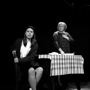 Two women on a stage, one sitting and the other standing behind a table with a checkered tablecloth.