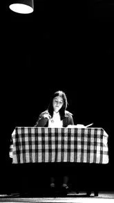 A woman sitting at a checkered tablecloth table, underneath a hanging light in a dark room.