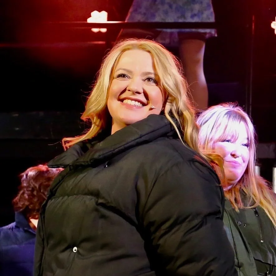 A woman with blonde hair smiling, wearing a black jacket, in a lively setting with other people around.