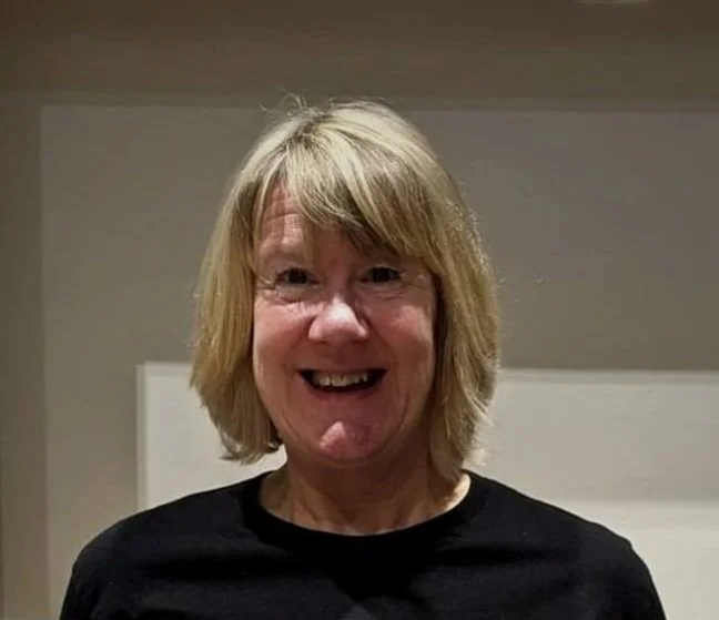 A woman with blonde hair smiling, standing indoors in front of a white wall.