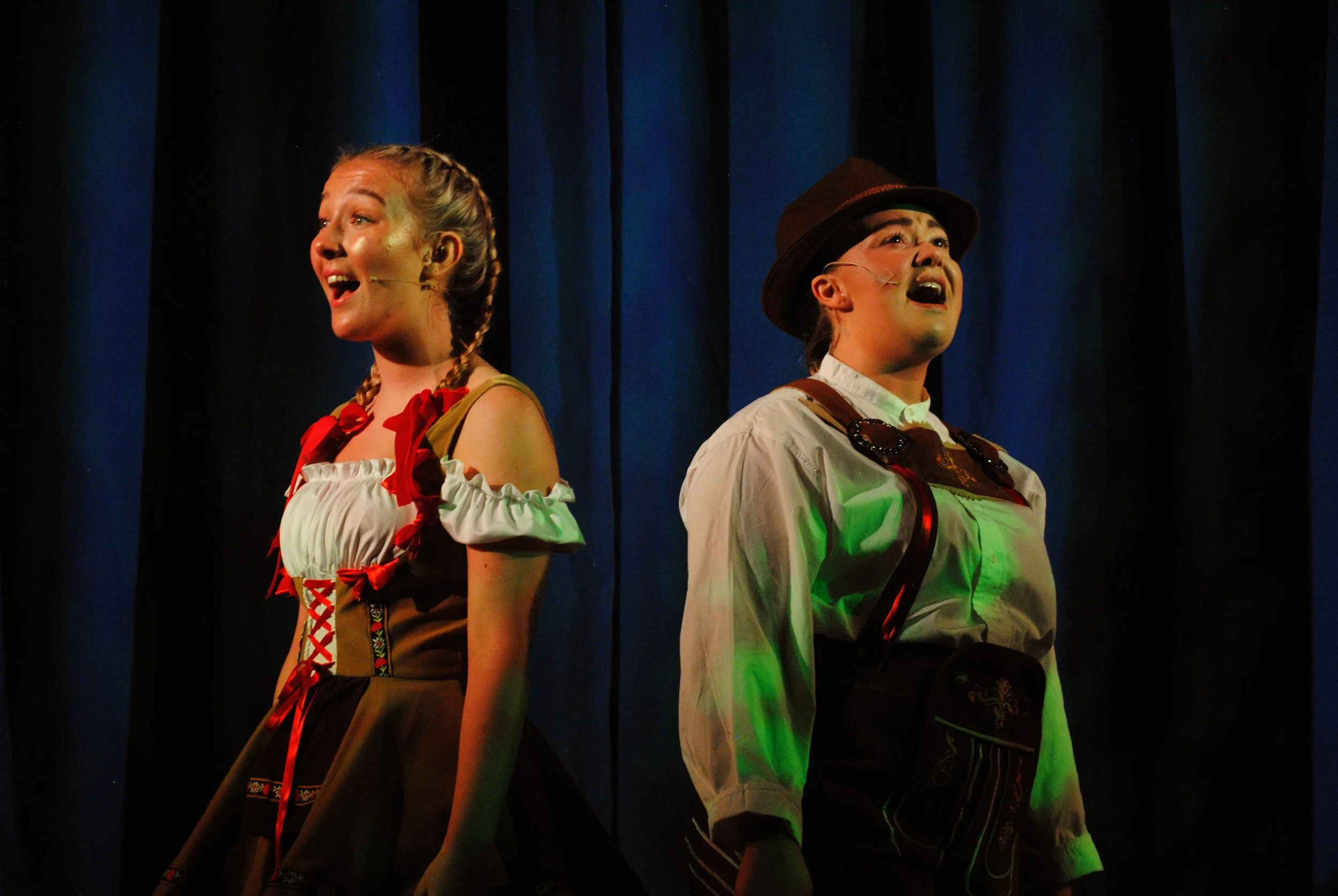 Two performers dressed in traditional Bavarian costumes singing on stage with blue curtains in the background.
