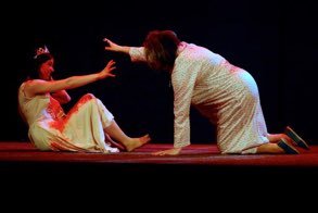 Two women dressed in traditional clothing on stage, one is sitting on the floor reaching out, and the other is kneeling and leaning forward, reaching out toward the sitting woman.