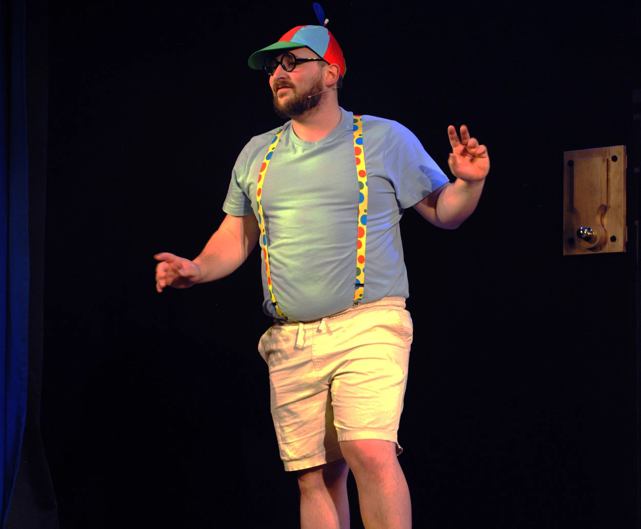 A man with glasses, a beard, wearing a colorful cap, gray T-shirt, and shorts, standing on stage with a dark background, gesturing with his hands.
