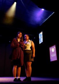 Two young women standing on a stage with colored lights and screens behind them.