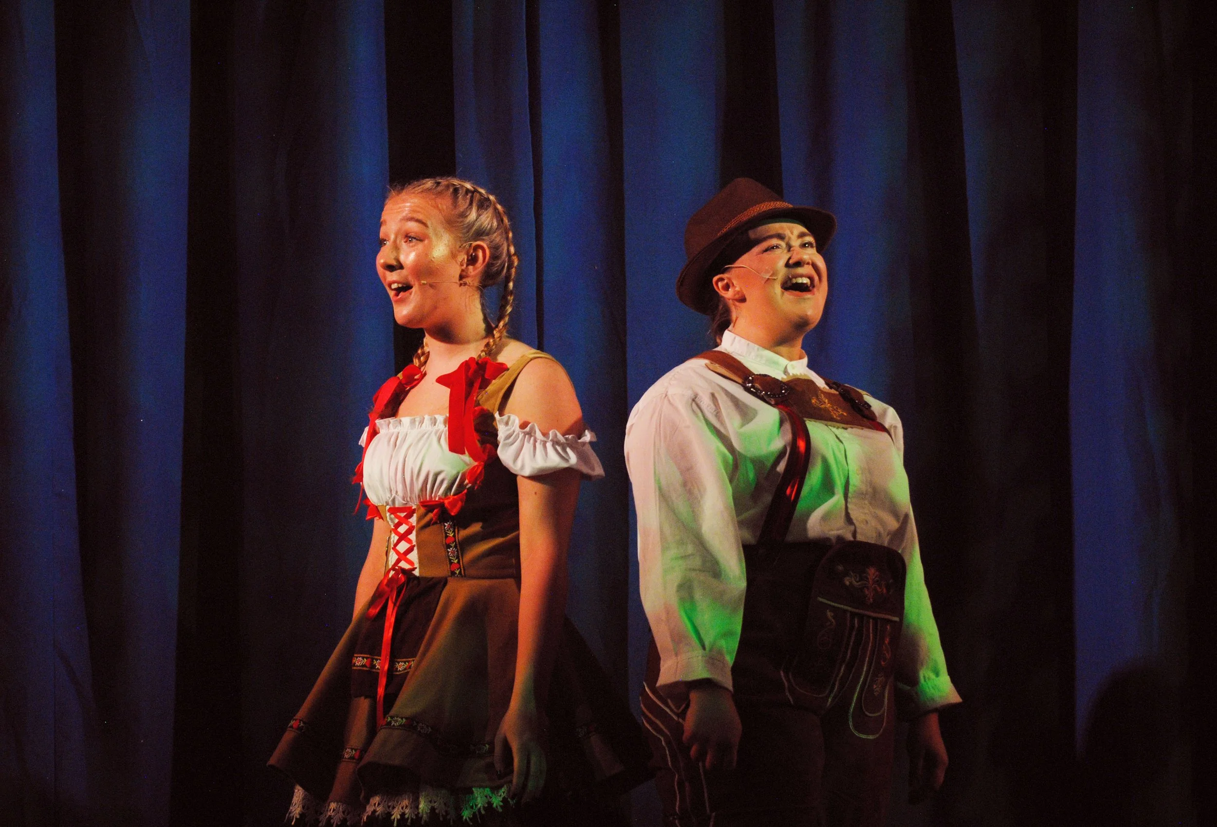 Two young girls performing on stage, wearing costumes and singing, with a dark blue curtain in the background.