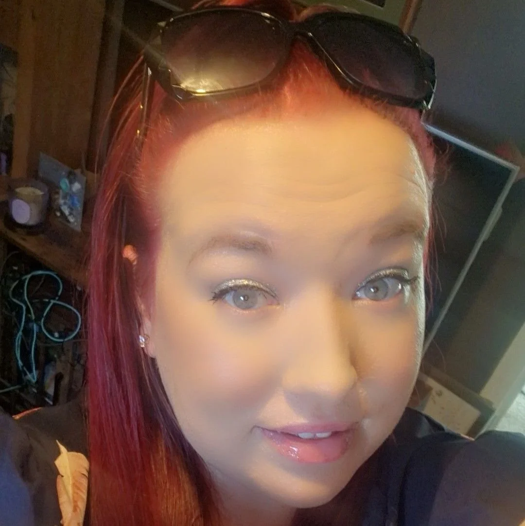 A woman with red hair, wearing sunglasses on her head, smiling, with a TV and various items in the background.