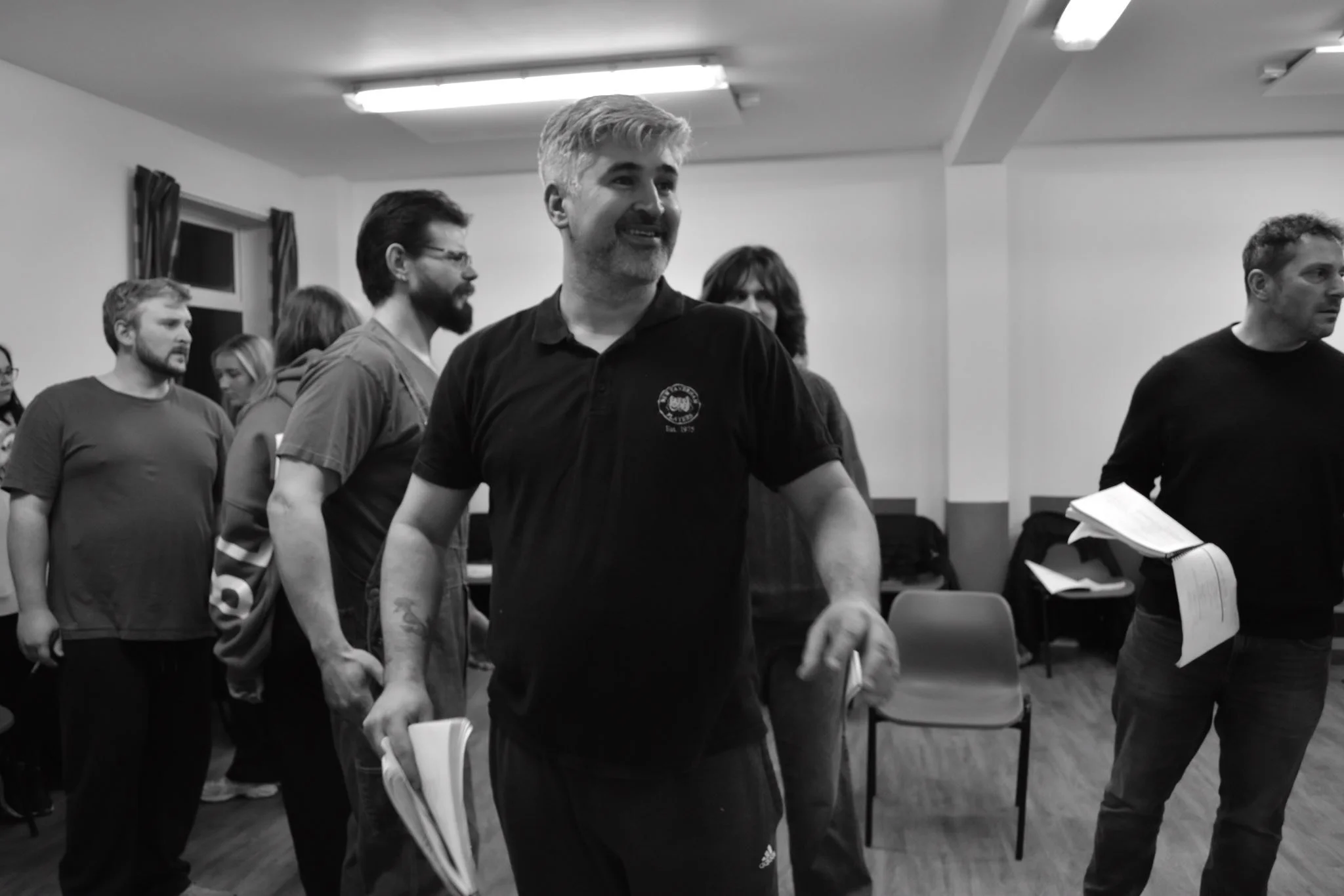 A group of people standing in a room, some holding papers, with a man in the foreground smiling and holding papers, in a casual gathering or meeting setting.
