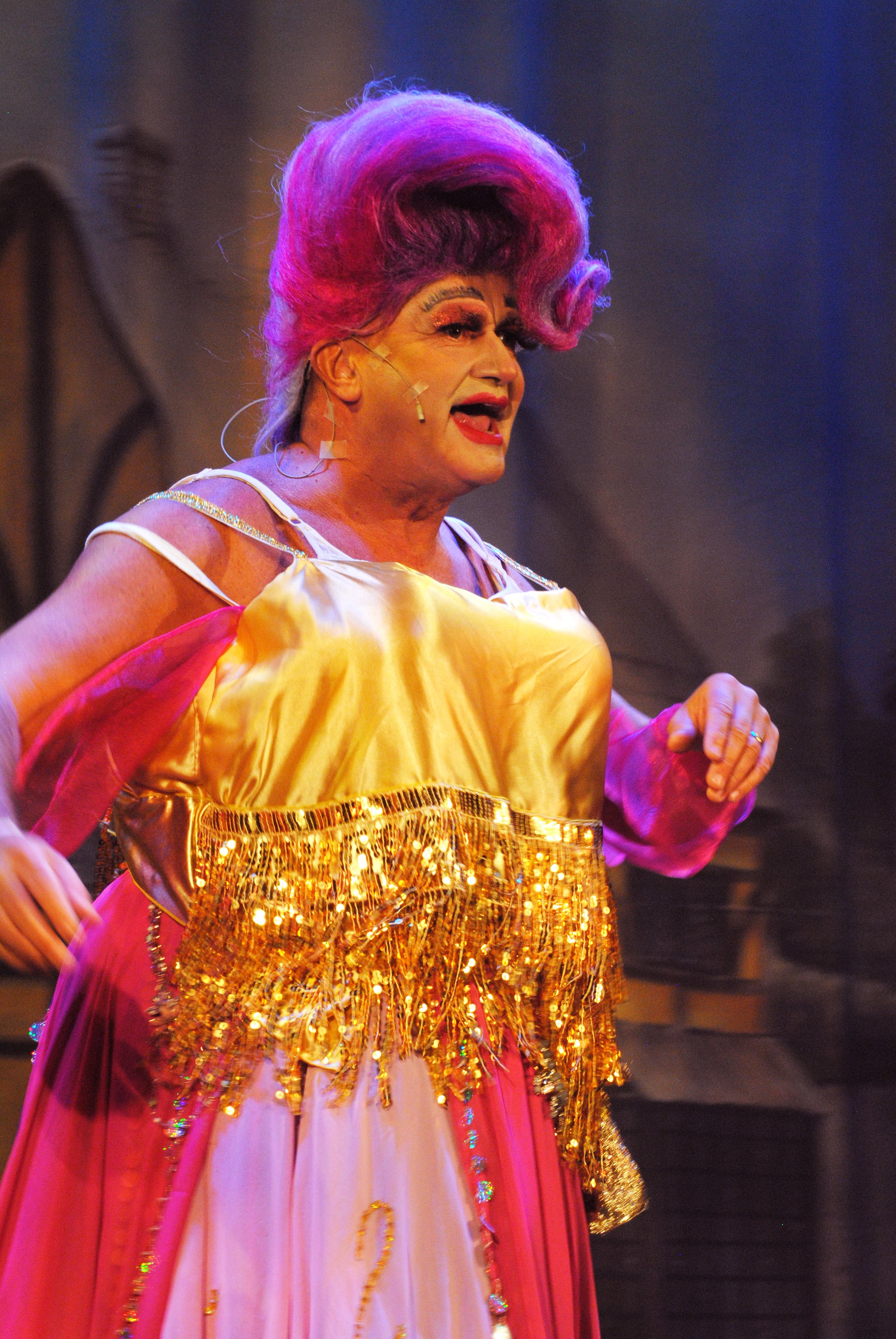 A performer with elaborate pink and purple hair, wearing a shiny gold and pink costume with gold fringe, singing on stage.