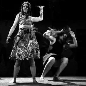 Two women performing on stage, one standing and the other crouching with her head tilted back and hand on her head, in black and white.
