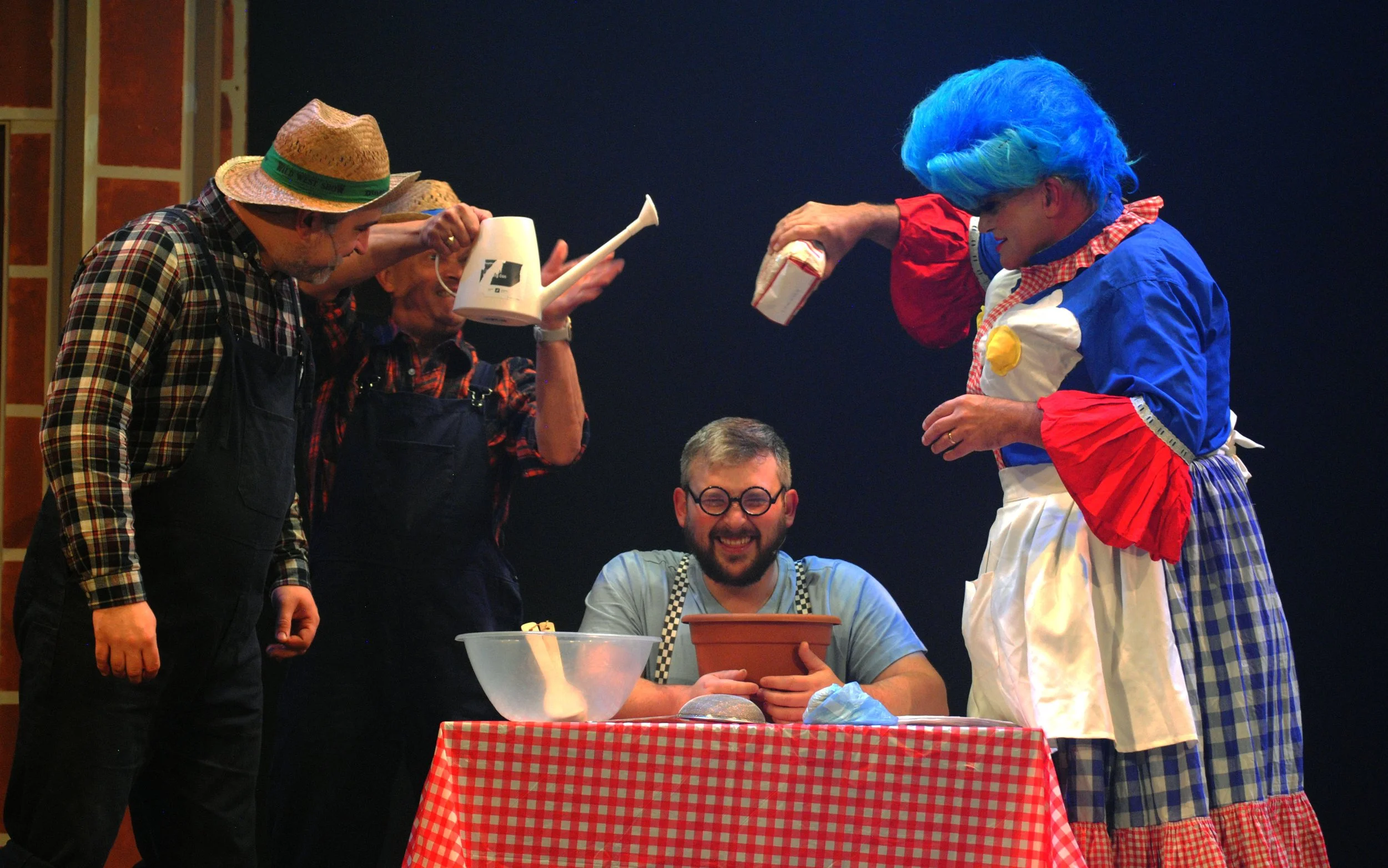 People dressed as farmers and a clown gathered around a table for a stage performance, with one person smiling while holding an object, and others holding watering can, bags, and watering can, with a checkered tablecloth.