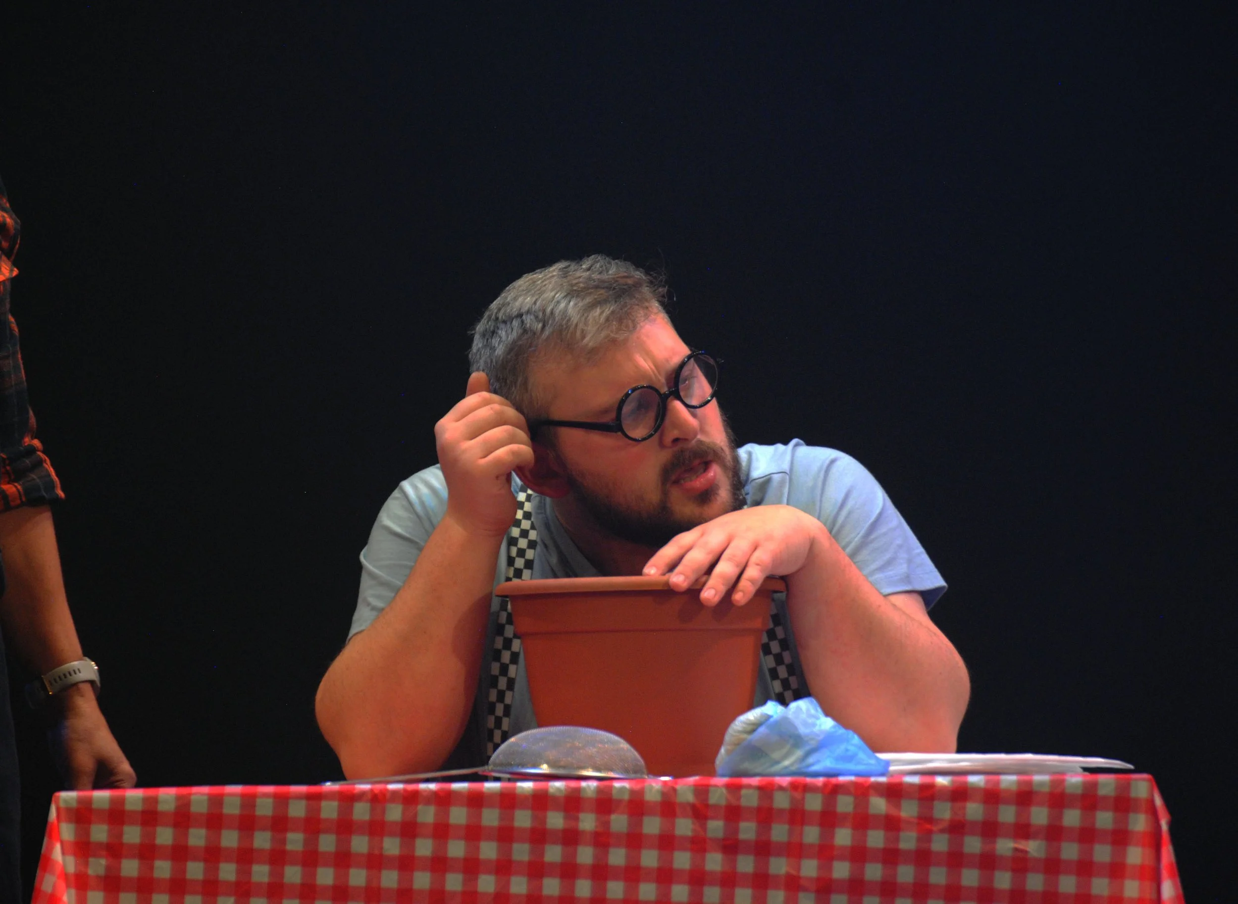 A man with glasses, gray hair, and beard, resting his head on his hand, appears confused or deep in thought, sitting at a table with a red and white checkered tablecloth, a terracotta pot, a blue face mask, and some papers.