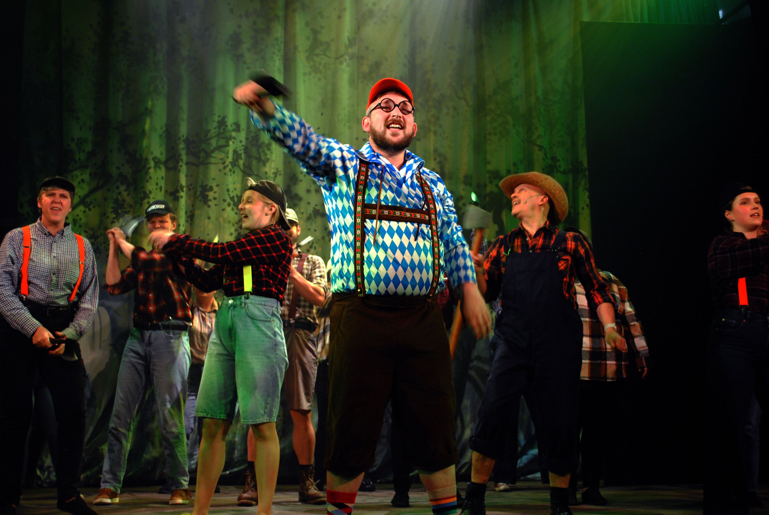 A theatrical performance with actors dressed in rural, traditional German or Alpine costumes, including checkered shirts, suspenders, lederhosen, and a woman in a plaid dress. The background features a forest scene with green lighting, and the actors