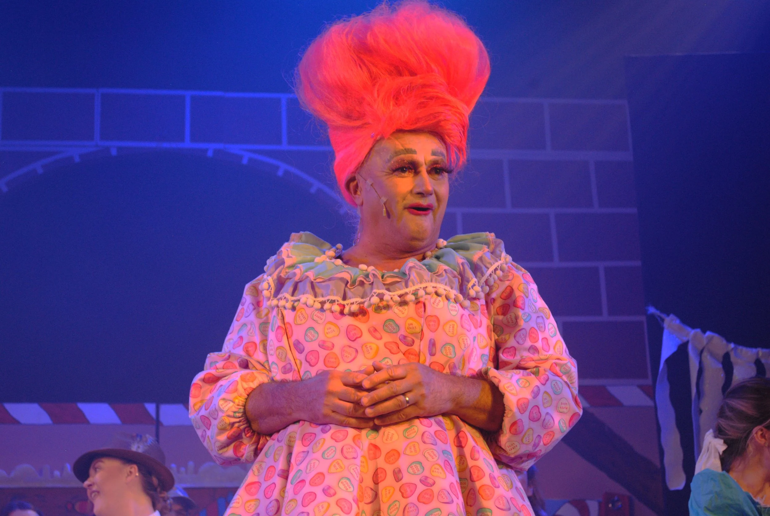 An actor with exaggerated pink hair and makeup dressed in a colorful costume, standing on stage during a theatrical performance.