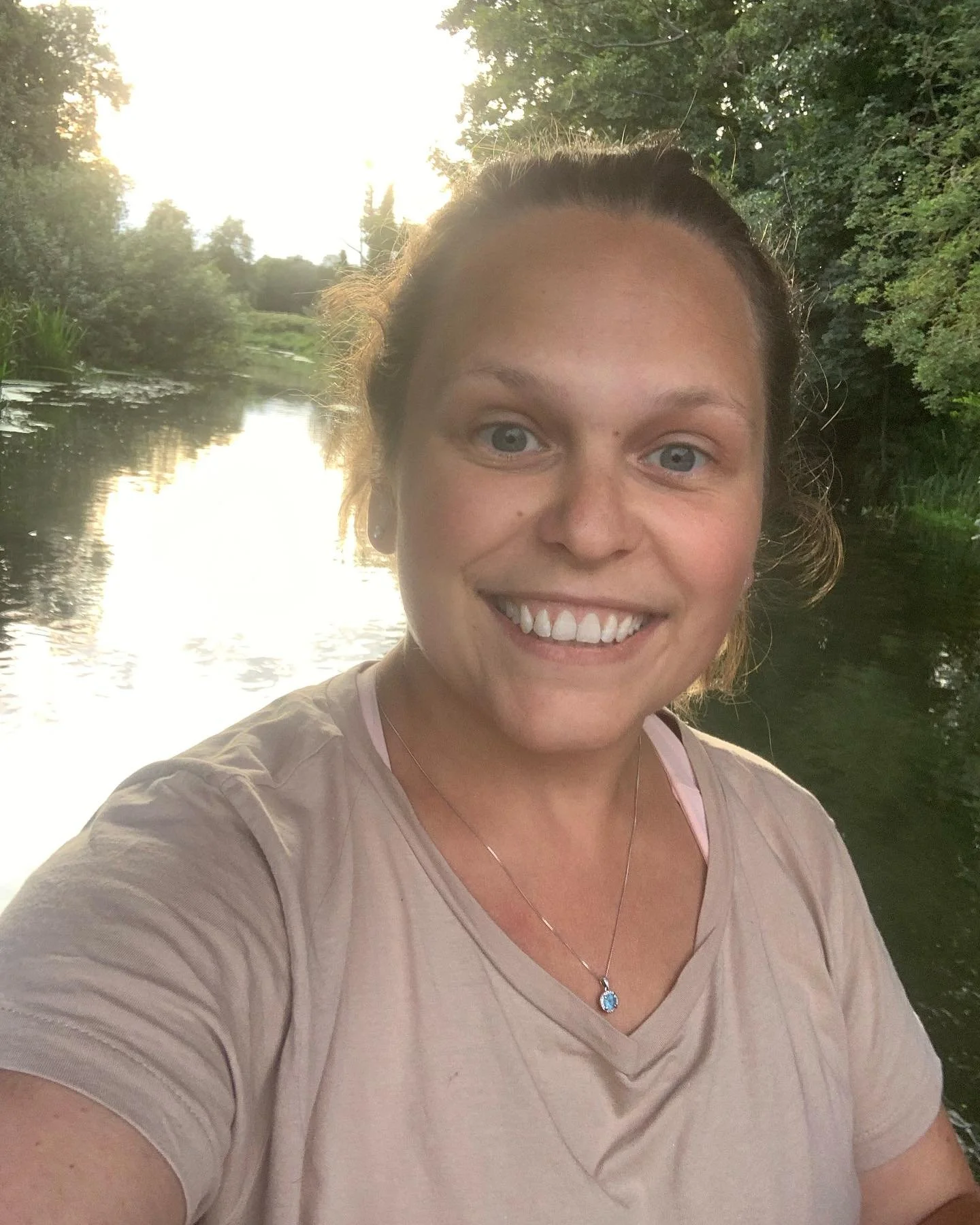 A woman smiling for a selfie outdoors near a river with trees and greenery in the background, and the sun setting in the distance.