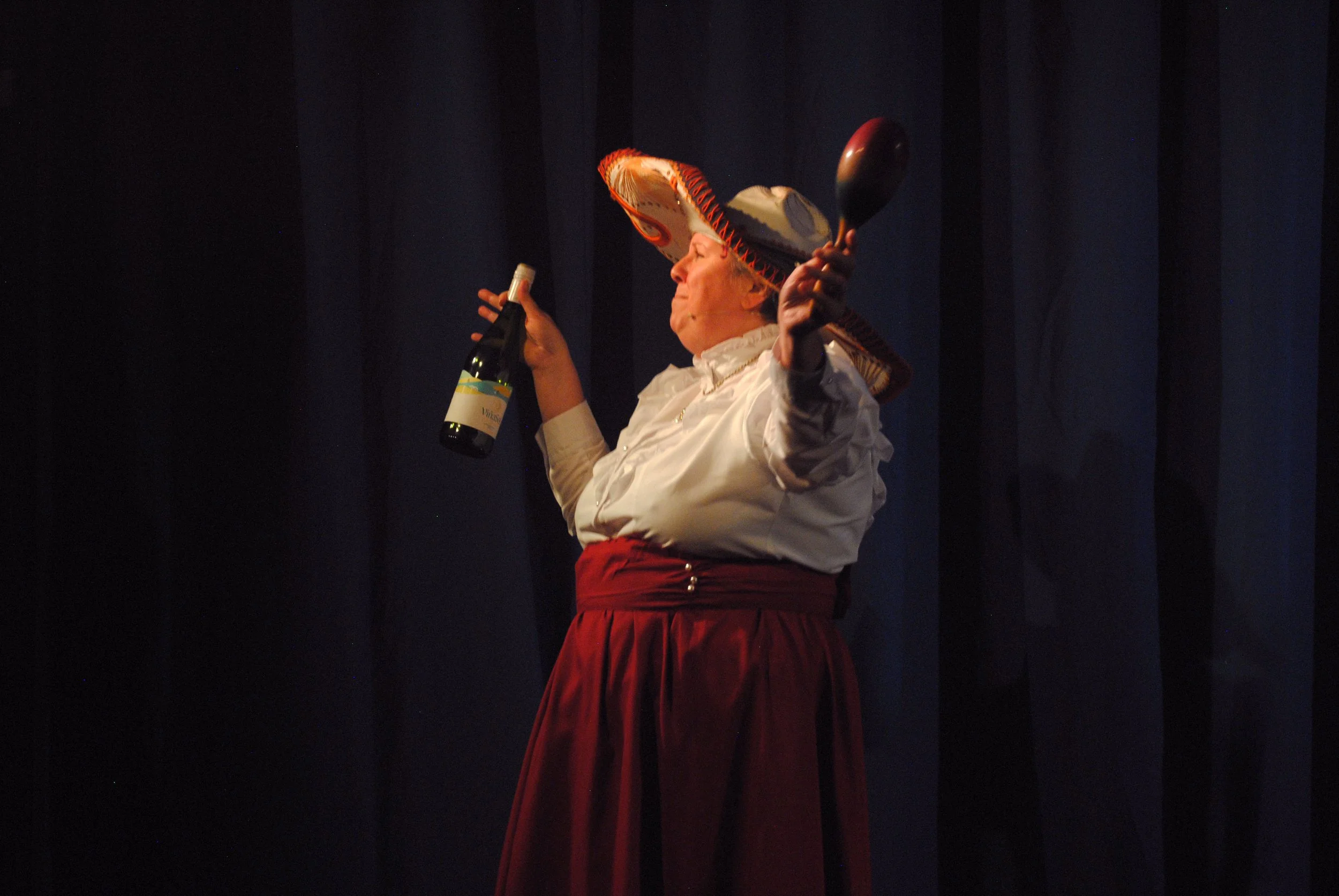 A woman wearing a traditional Mexican sombrero, red skirt, and white blouse holding a bottle of wine and a spoon, on a stage with a dark curtain background.