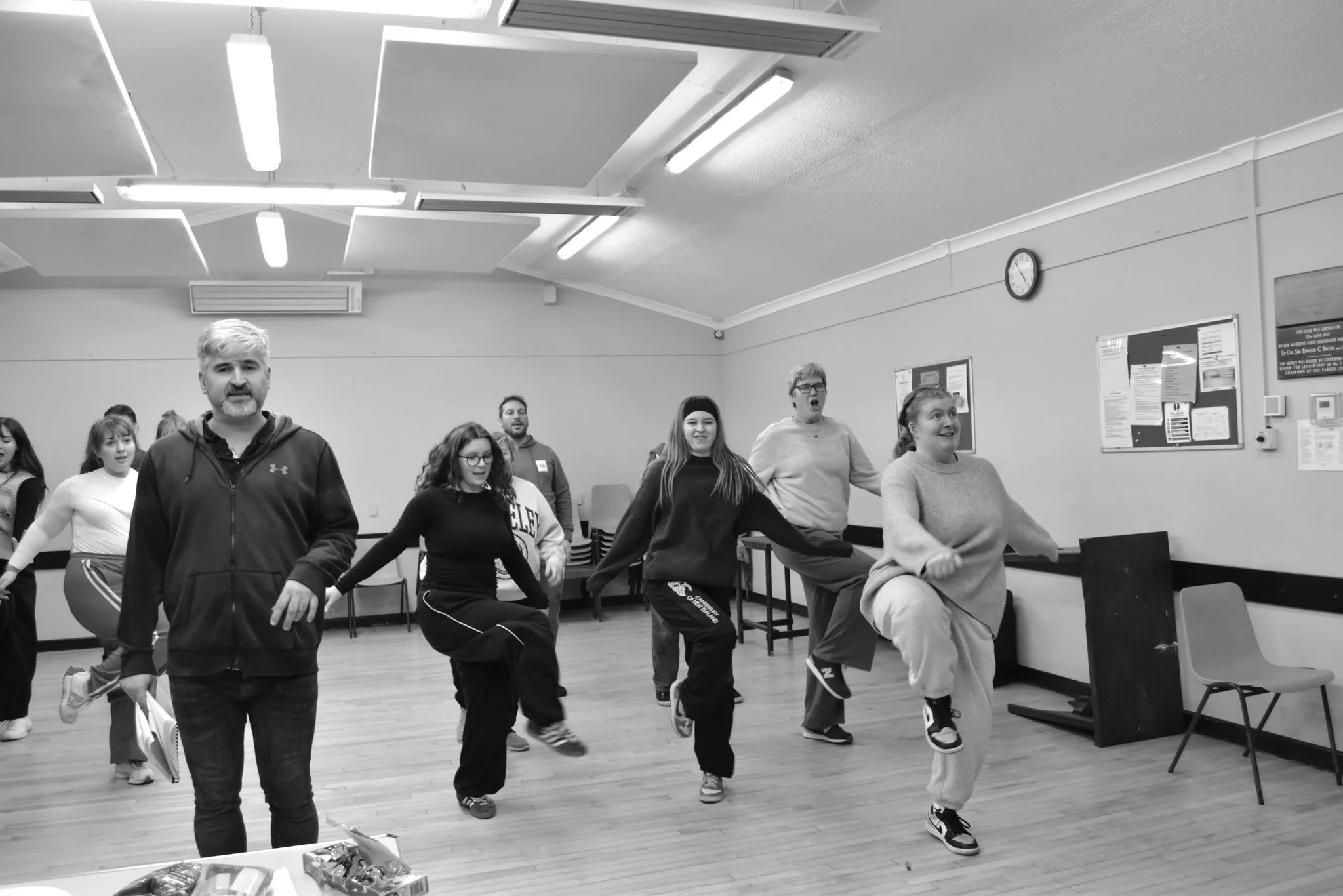 People participating in a dance or exercise class inside a room with wooden floors, ceiling lights, and a clock on the wall.