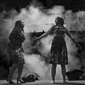 Two women on stage, surrounded by smoke, one wearing a zebra-patterned dress and the other wearing a floral dress, appears to be performing or in conversation.
