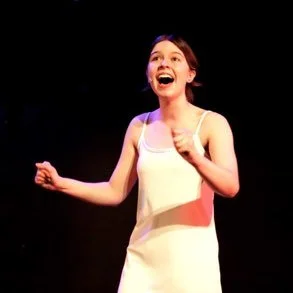 A woman with short brown hair, wearing a white sleeveless dress, smiling and gesturing with her right hand, standing on a stage with a black background.