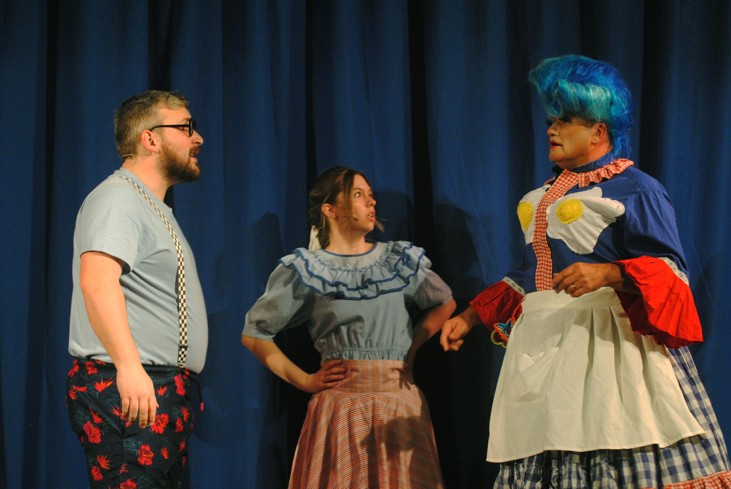 Three people on stage in a theatrical performance, with a man and a woman facing a clown with blue hair, in front of a blue curtain.